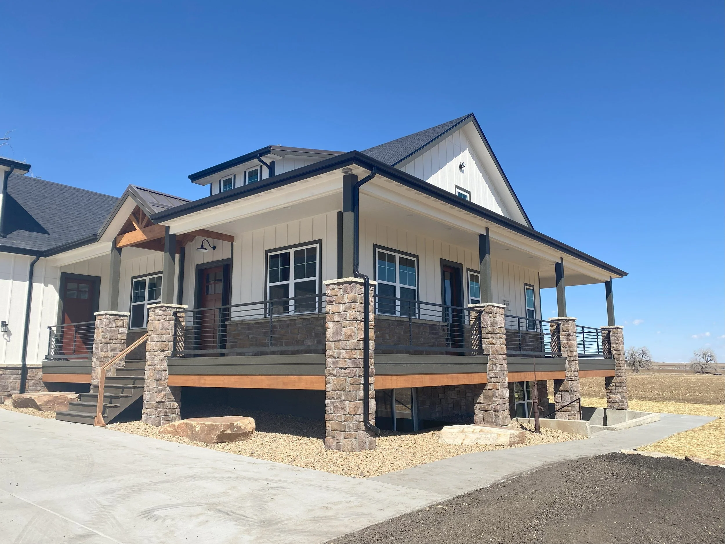 New modern house with white siding, black accents, and a large porch with stone pillars and black railing, under a clear blue sky.