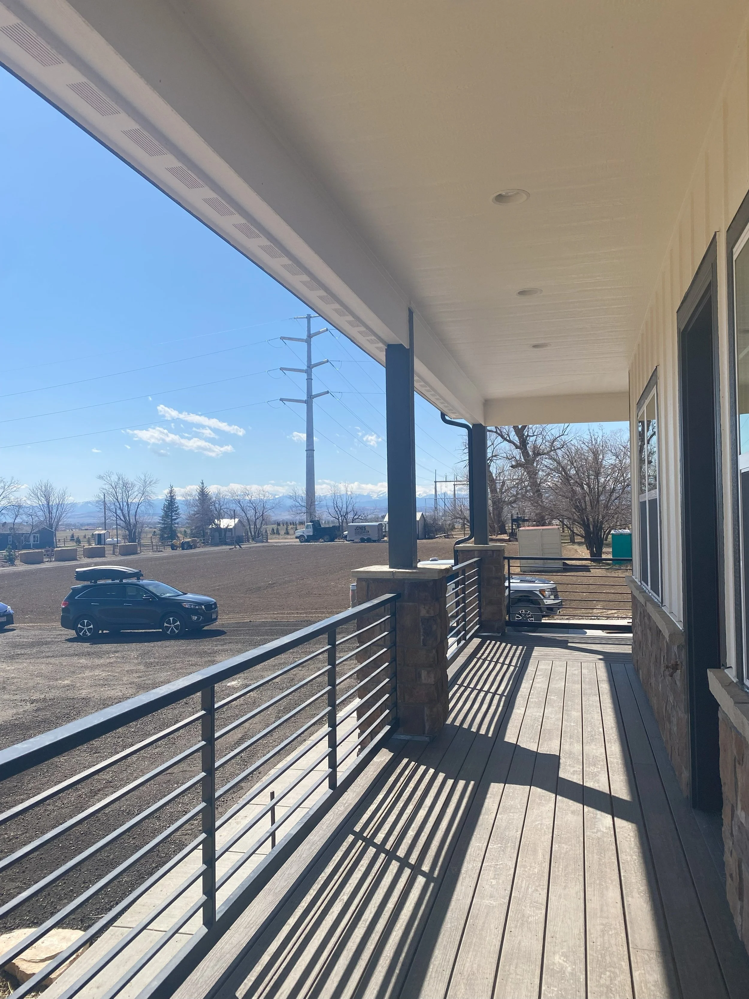 View from a porch showing a dirt parking lot with two black cars and a truck, a field, and bare trees with mountains in the distance under a clear blue sky.