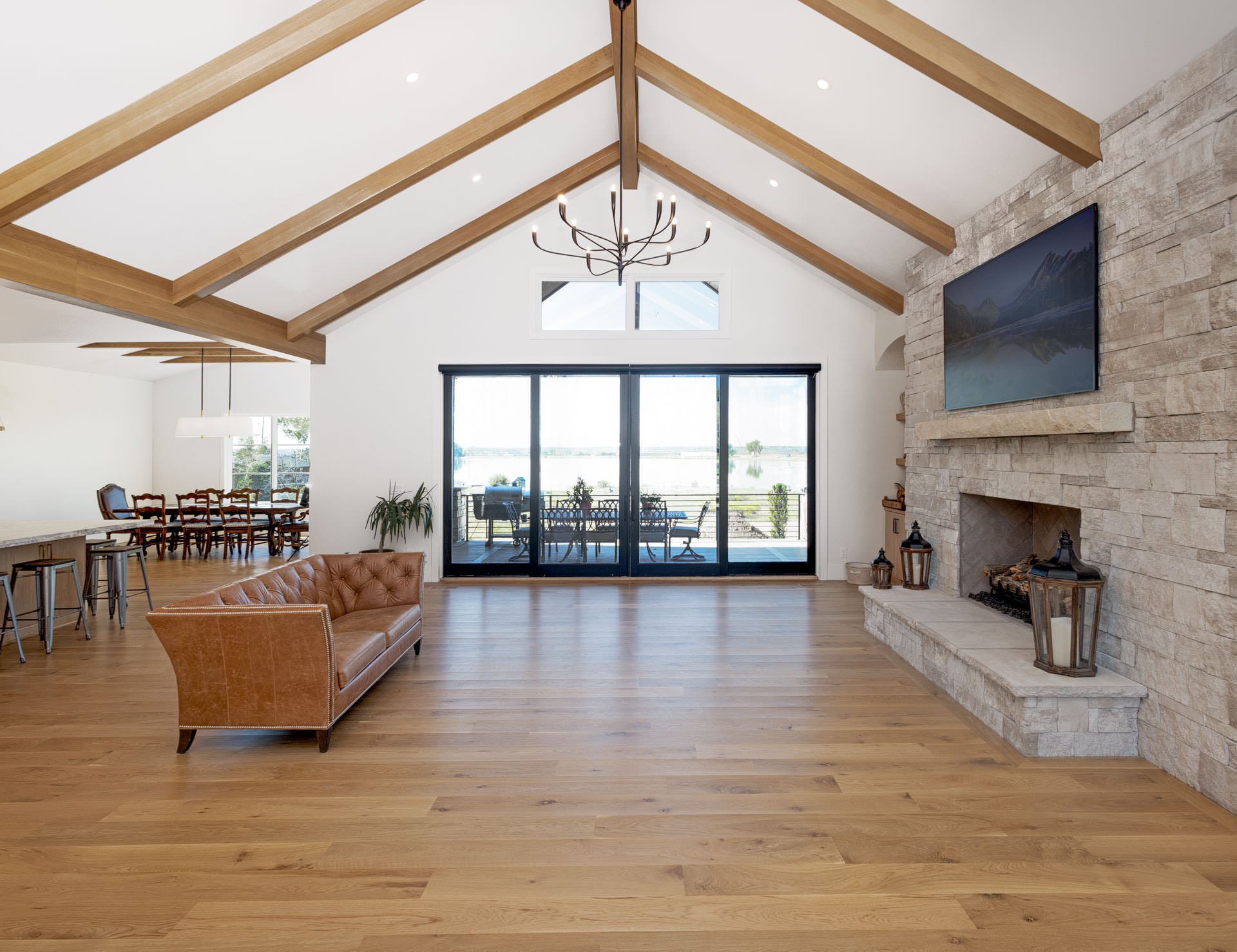 Spacious living room with a high vaulted ceiling with wooden beams, a large stone fireplace with a mounted flat-screen TV, sliding glass doors leading to a balcony with outdoor seating, and a view of a water body.