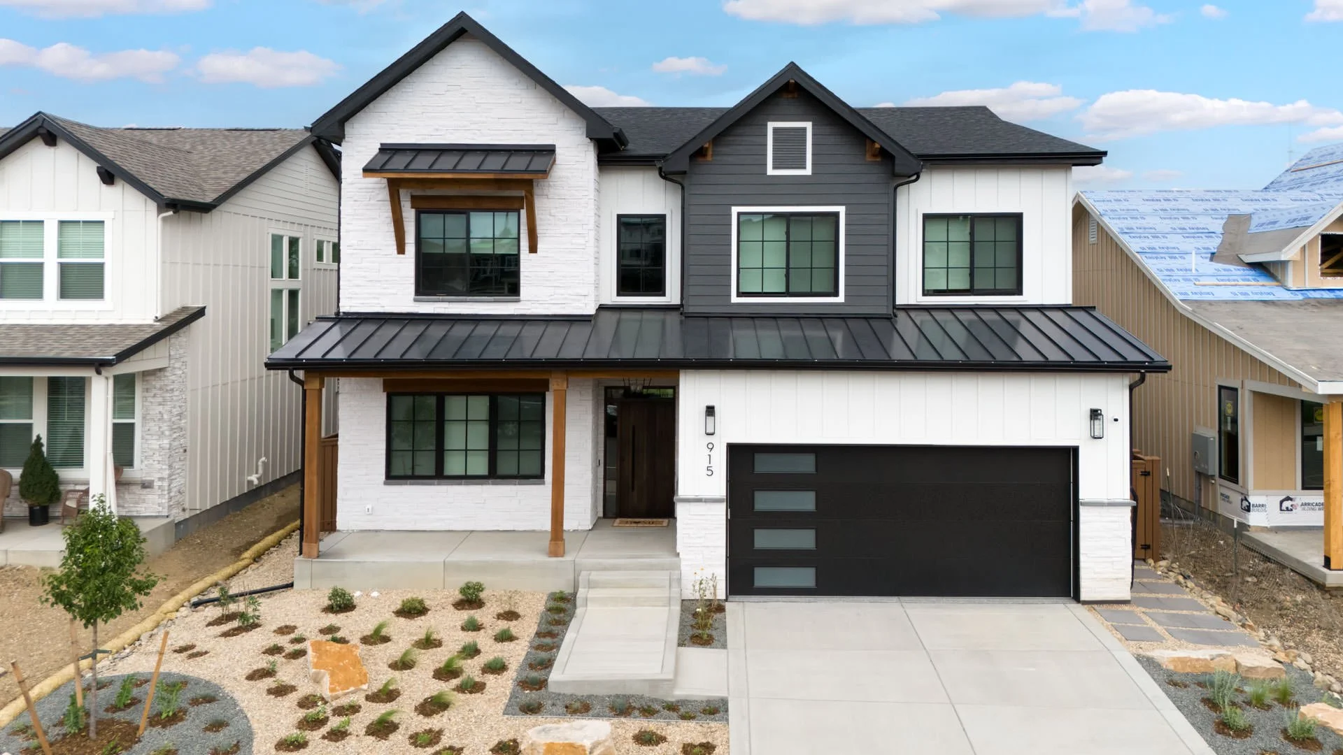 Front view of a modern two-story house with a black metal roof, white brick facade, black window frames, and a black front door. The house features a two-car garage with a black door and a small porch with wooden posts. The front yard has minimal landscaping with small plants and gravel pathways, and neighboring houses are under construction.