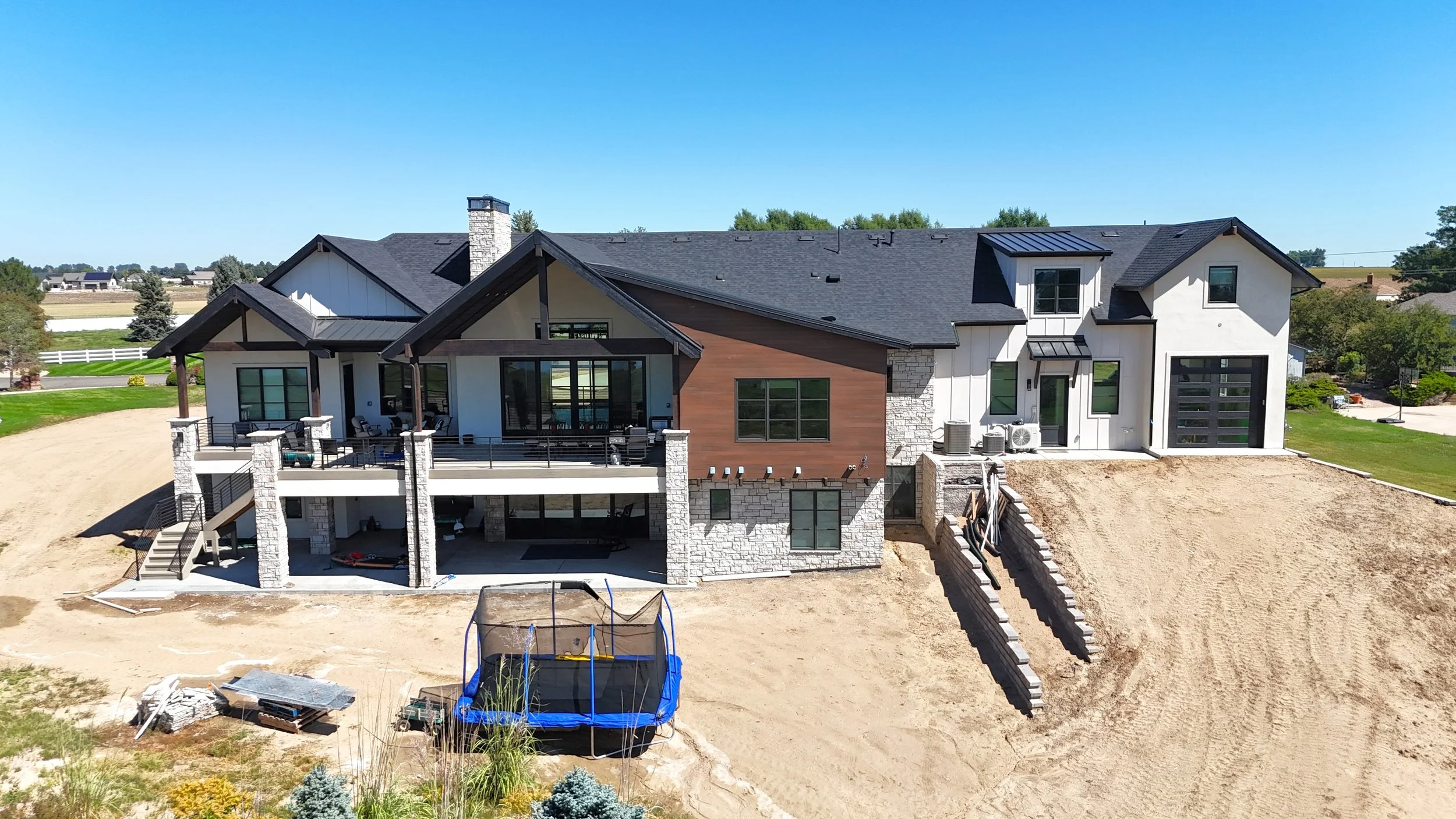 A modern two-story house under construction with a large balcony, outdoor seating area, and multiple windows, situated on a dirt lot with some construction equipment and a trampoline in the backyard.