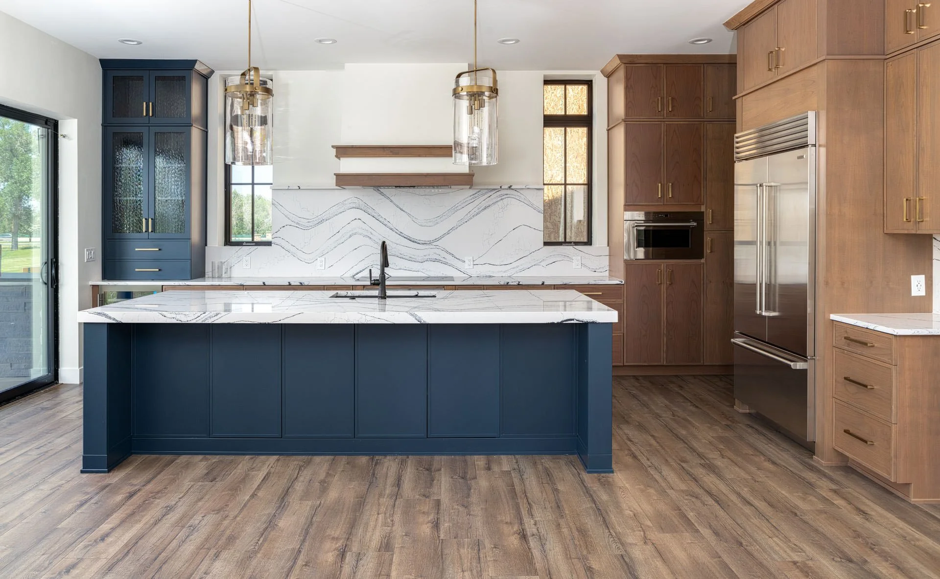 Modern kitchen with a large navy blue island, wooden cabinets, marble countertops, and stainless steel appliances, including a double-door refrigerator and wall oven. There are pendant lights hanging above the island and windows bringing in natural light.