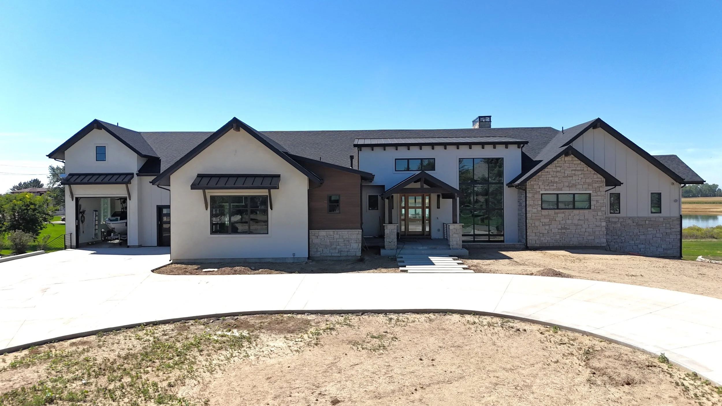 Newly constructed modern house with a combination of white, stone, and wood siding, large windows, and a front porch, situated in a spacious yard with a curved sidewalk.