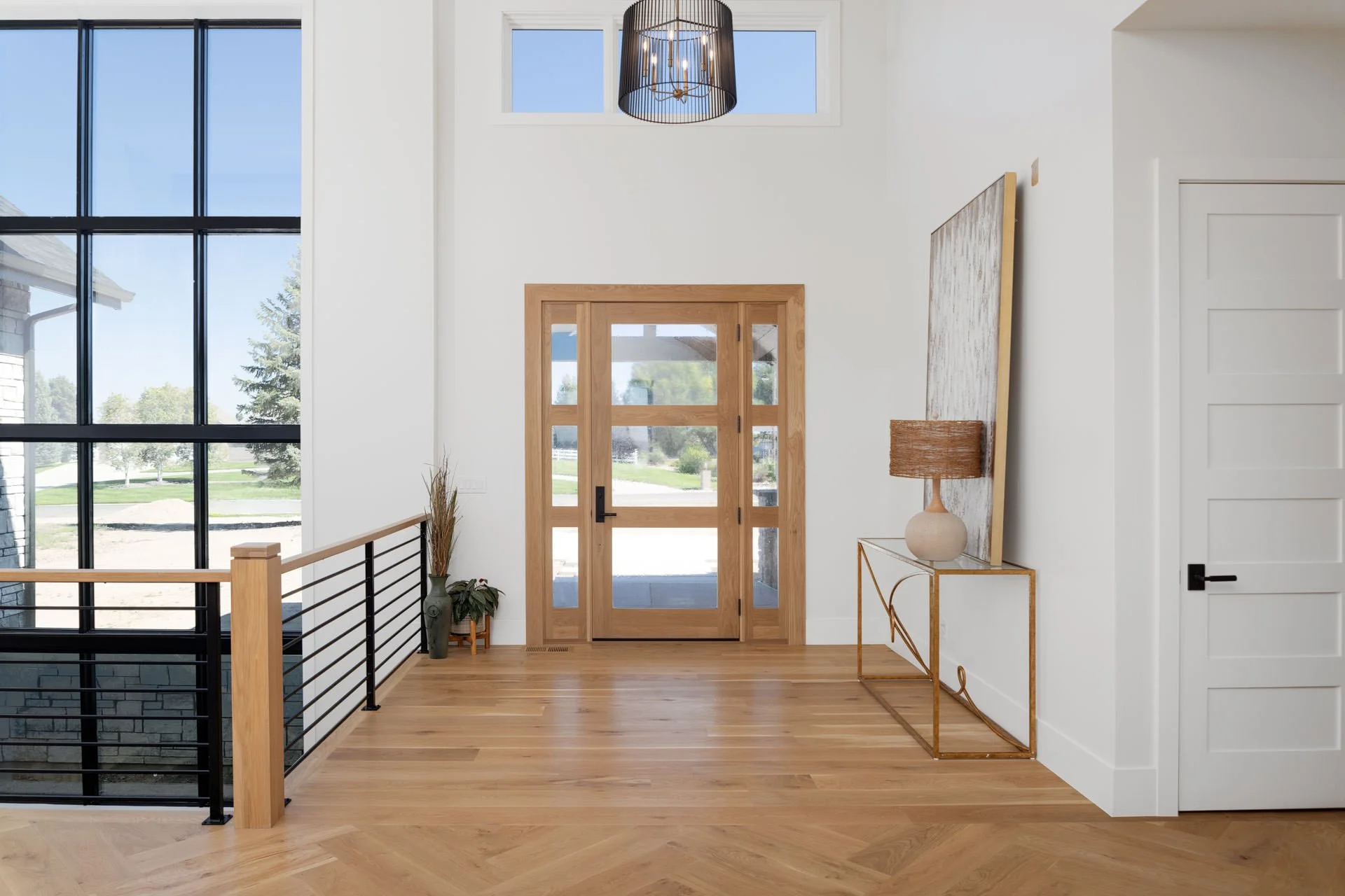 Entryway of a modern home with large windows, wooden door, and minimalist decor including a side table with a lamp and potted plants.