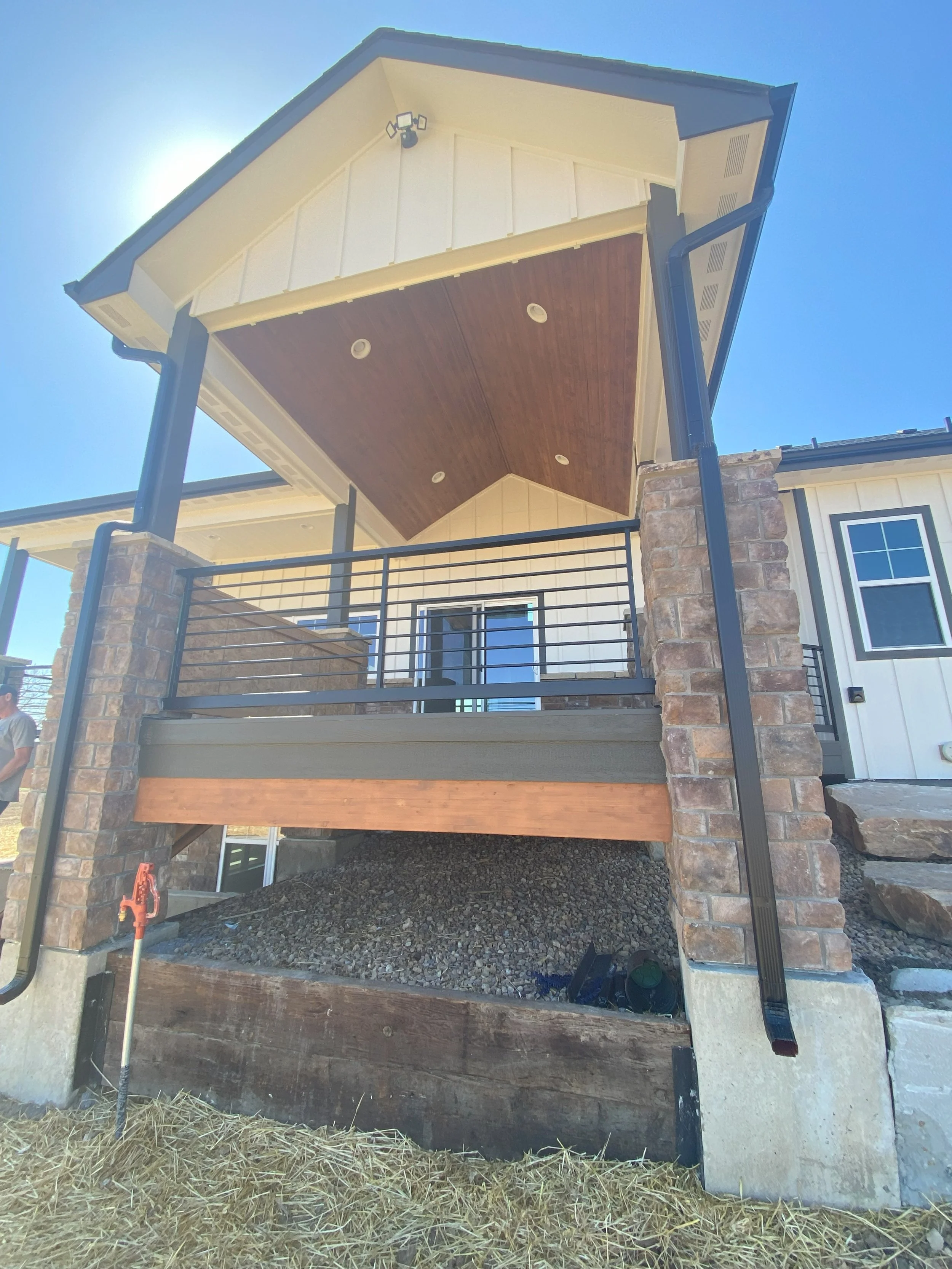 Front view of a house with a covered porch area featuring dark metal railings, brick columns, and a wooden ceiling. The house has white siding and black trim, with a visible window and door, set against a bright blue sky.