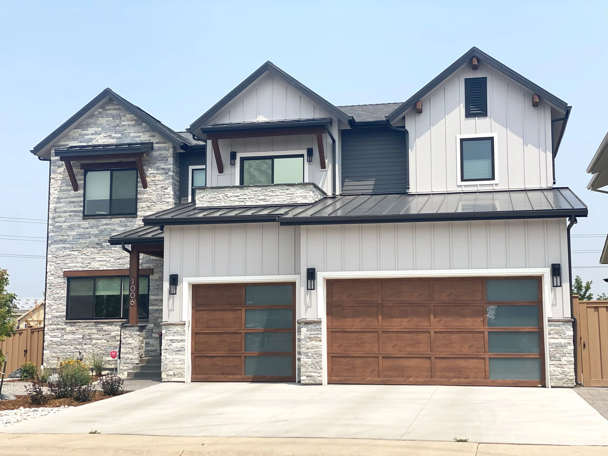 Modern multi-story house with a stone and white siding exterior, two wooden garage doors, and multiple windows under a partly cloudy sky.