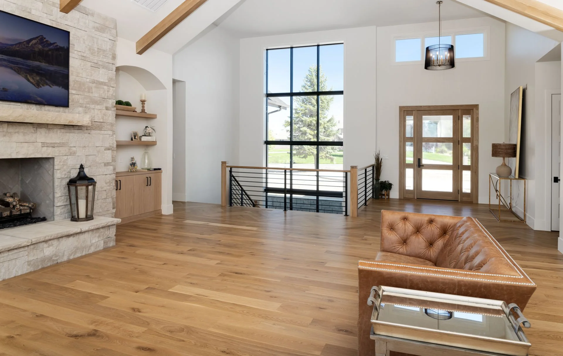 Bright living room with hardwood floors, a large stone fireplace, a leather sofa, and a wooden front door with glass panels. Floor-to-ceiling window and modern decor.