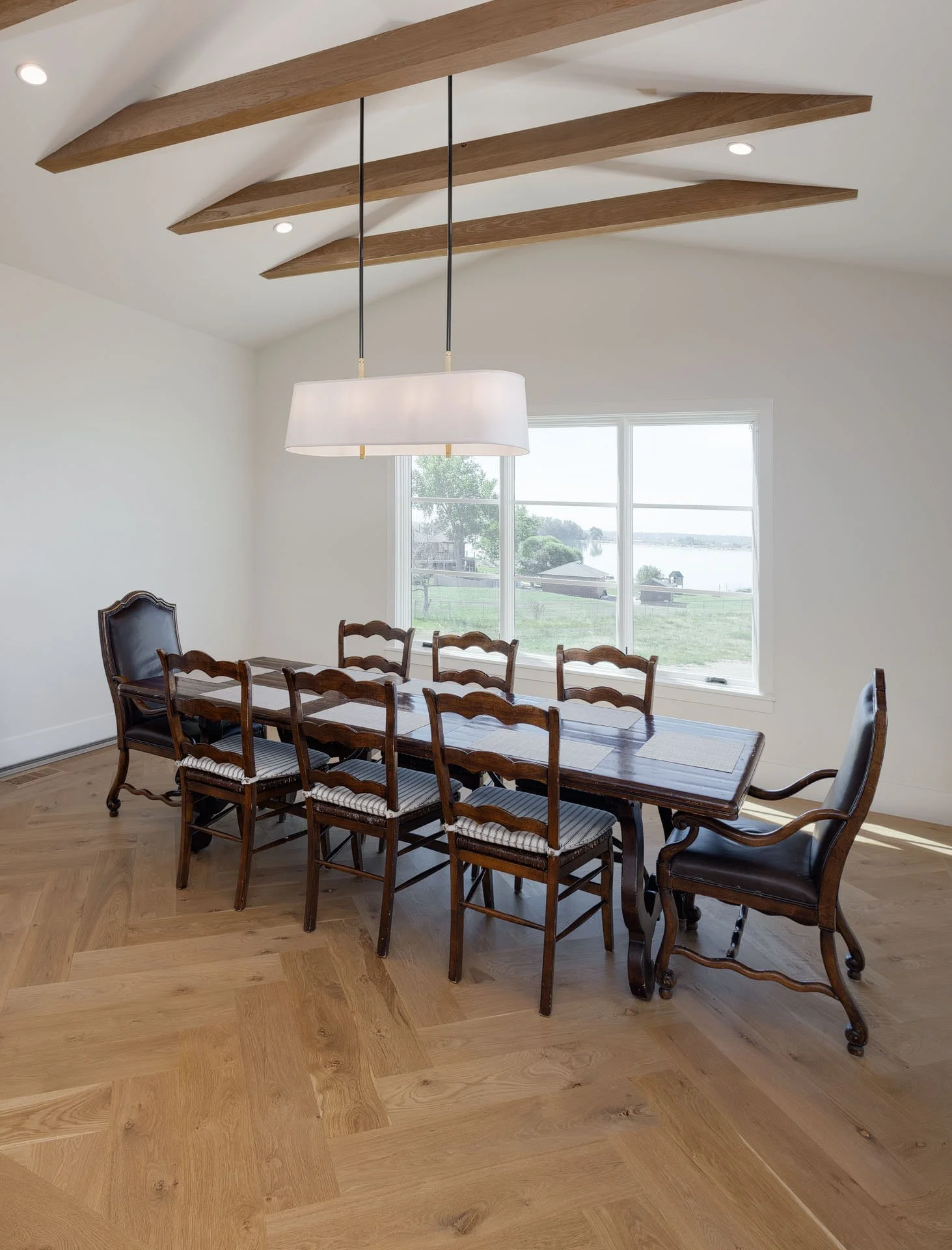 A dining room with a wooden table and eight chairs, two of which are armchairs, inside a space with white walls, skylight windows, and a large window overlooking a outdoor landscape with water, trees, and sky.