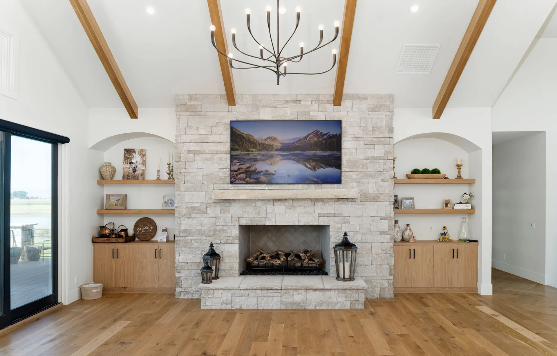 Living room with a stone fireplace, mounted television, wooden shelves with decorations, a chandelier, hardwood floors, and sliding glass door to the outside.