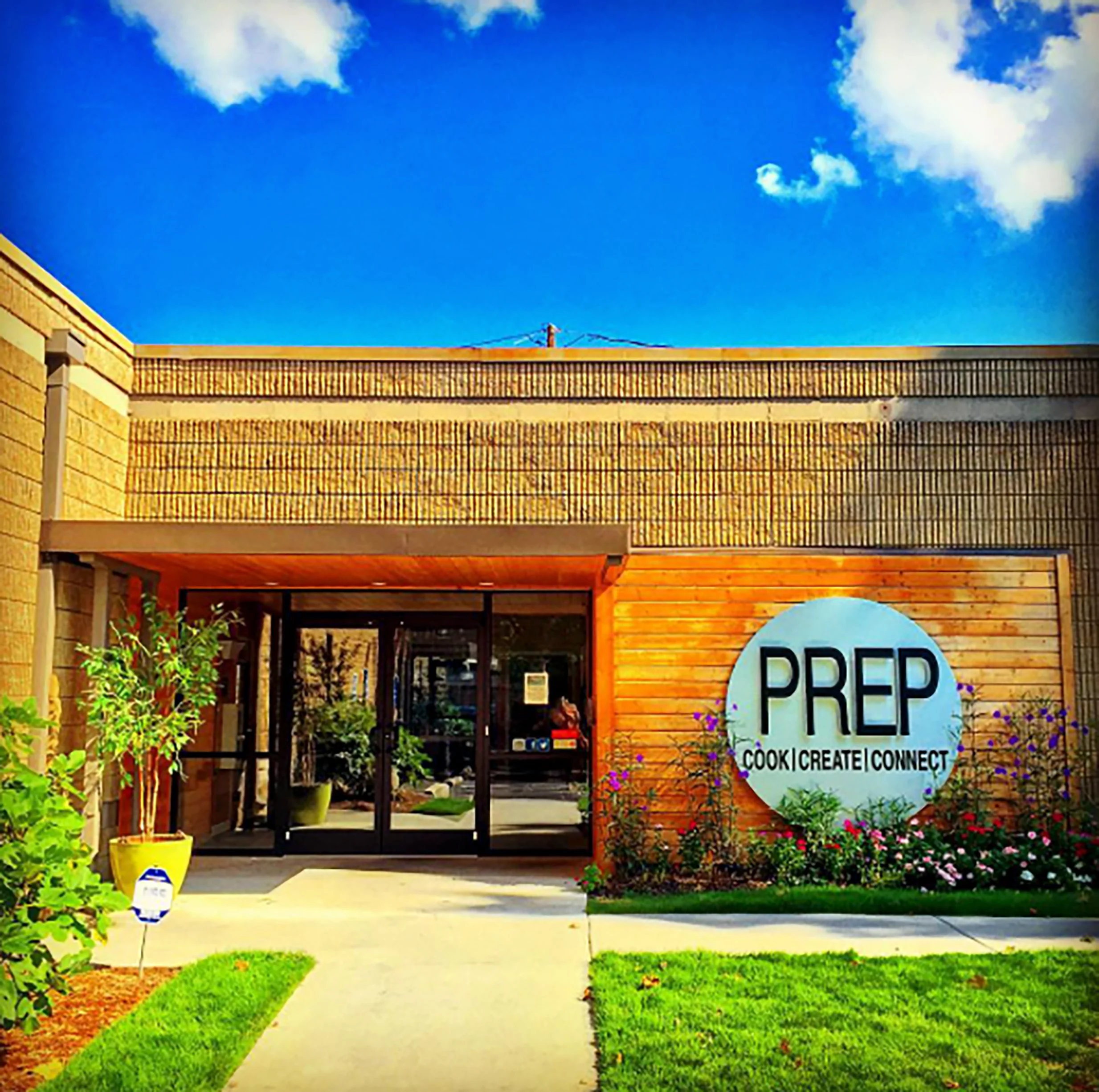 The entrance of a building with a round sign that reads 'PREP' and 'Cook | Create | Connect' with a garden and sidewalk in front.