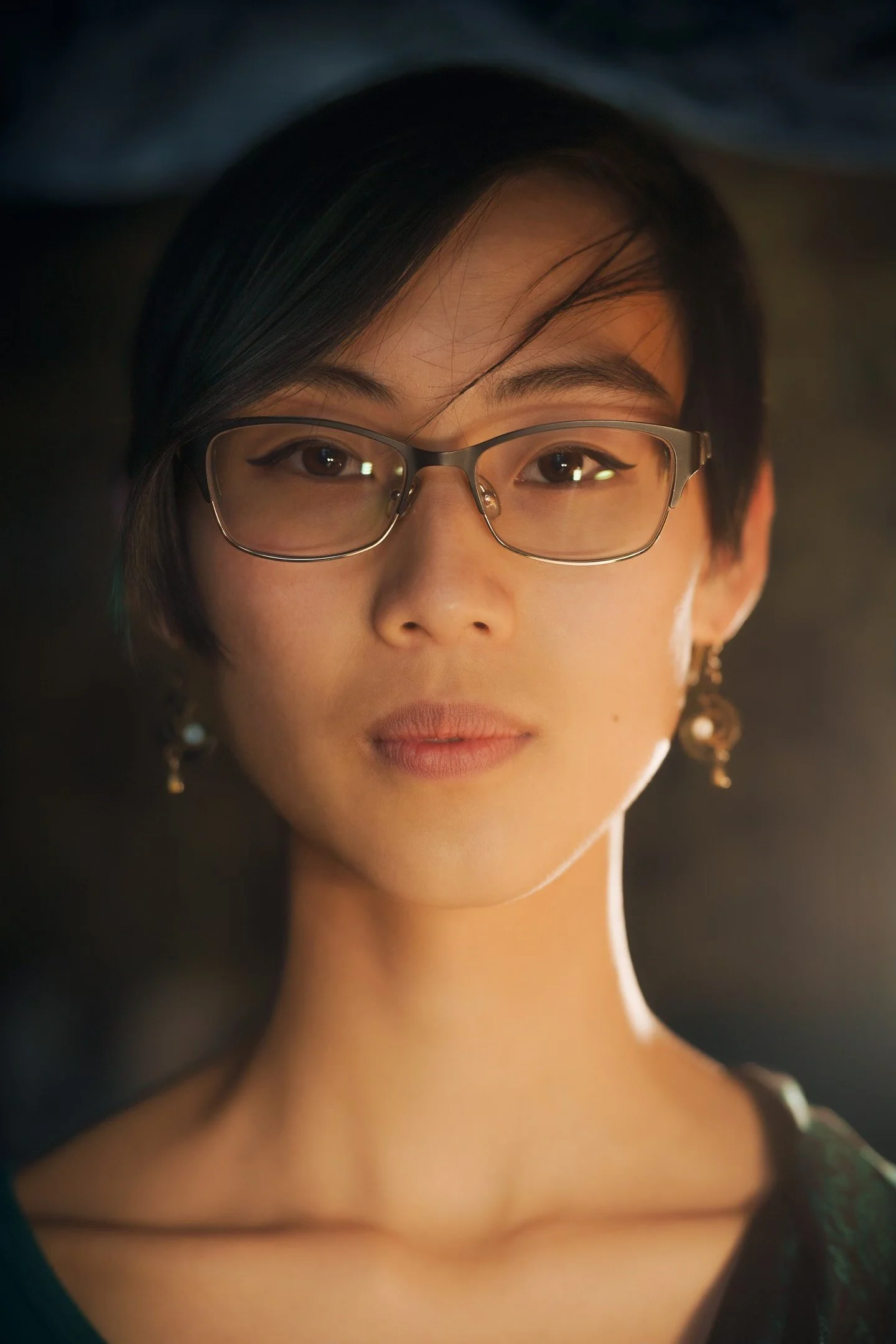 A close-up portrait of an Asian woman with short black hair, wearing glasses and earrings, looking directly at the camera with a neutral expression.
