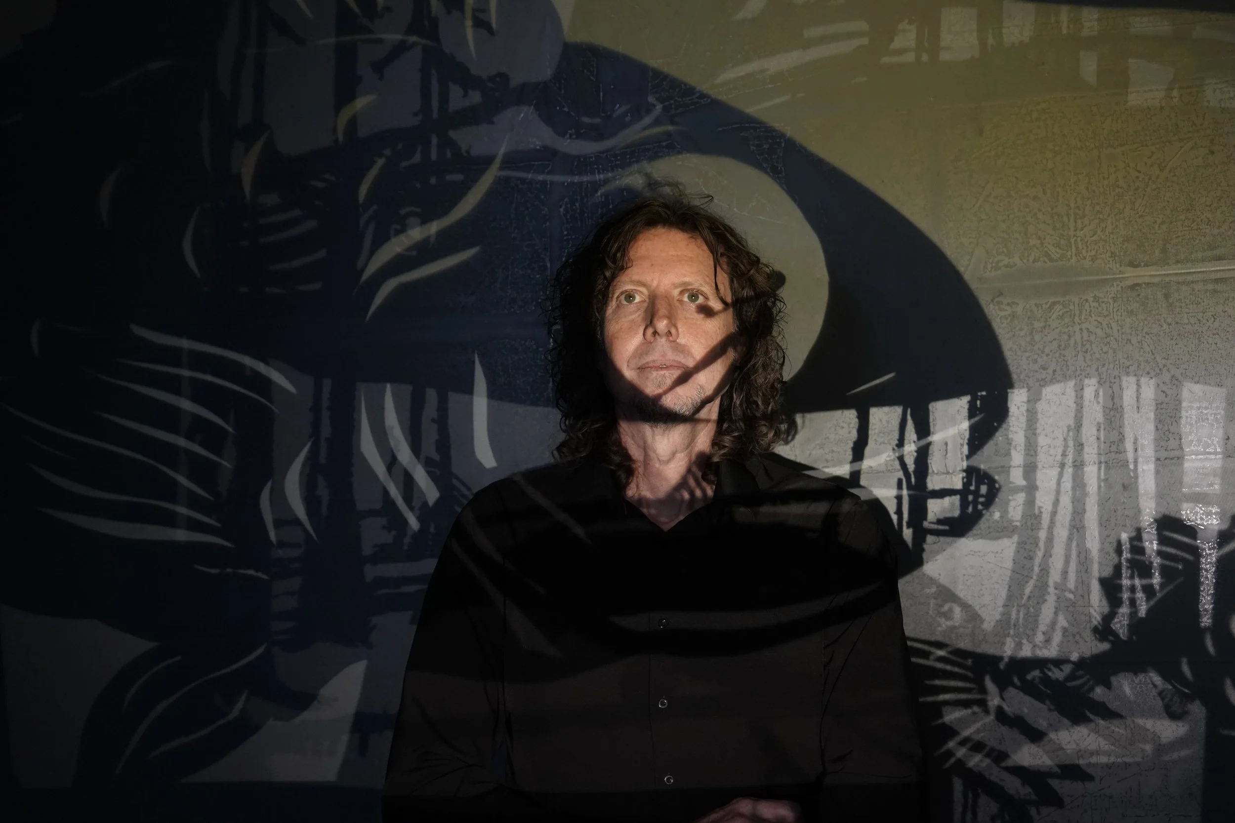 A man with curly hair standing in front of a dark background with a large, shadowed skeleton projection.