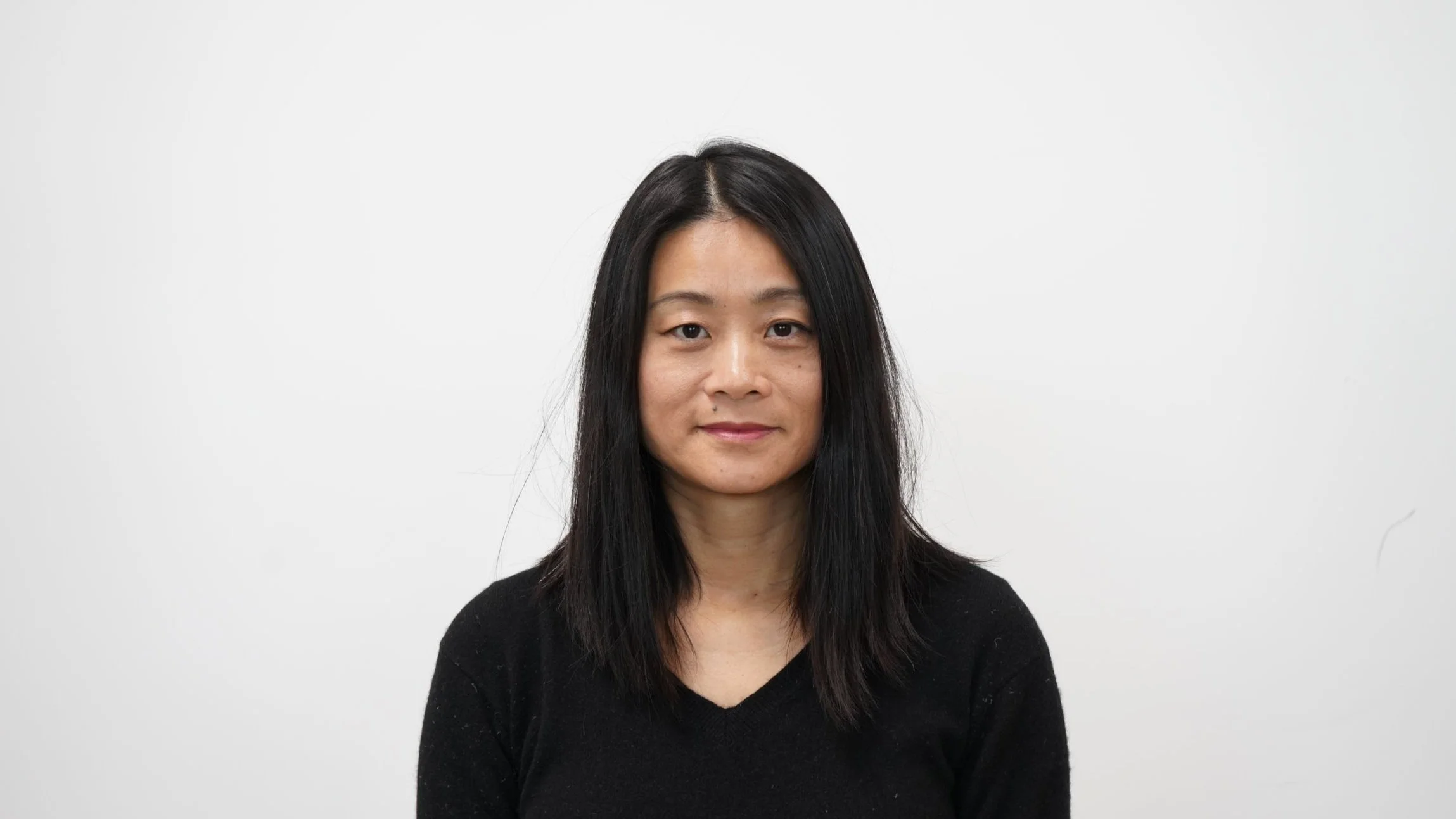 Portrait of a woman with straight, shoulder-length black hair wearing a black V-neck sweater, standing against a plain white background.