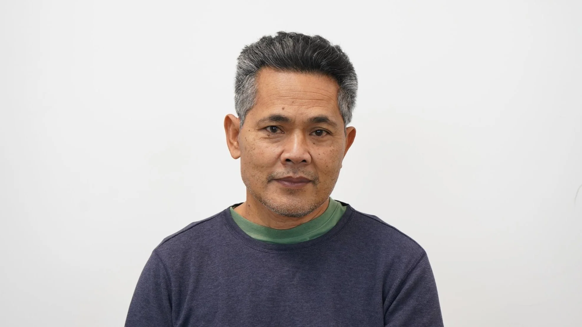 Portrait of an older man with salt and pepper hair, wearing a navy blue shirt over a green t-shirt, against a plain white background.