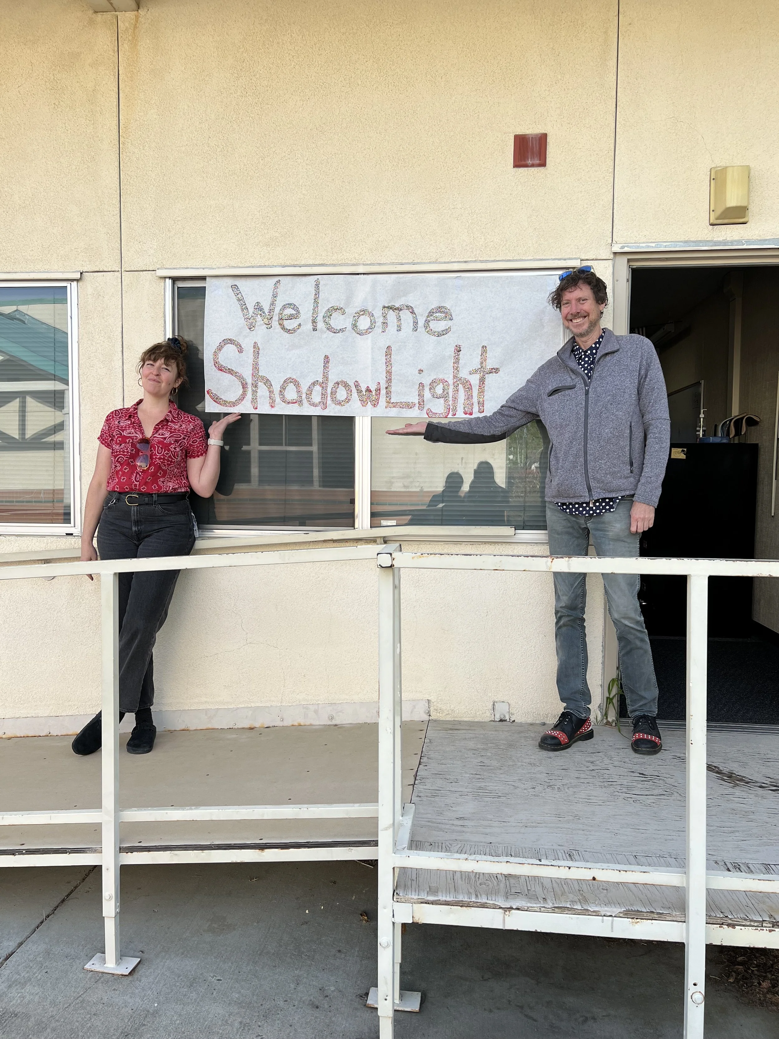 Two people standing on a small porch next to a window with a sign that says 'Welcome ShadowLight'. The woman on the left has curly hair and is wearing a red patterned shirt and black pants. The man on the right has curly hair, a beard, and is wearing a grey jacket, a polka dot shirt, jeans, and black shoes with red accents. He is smiling and pointing towards the sign.