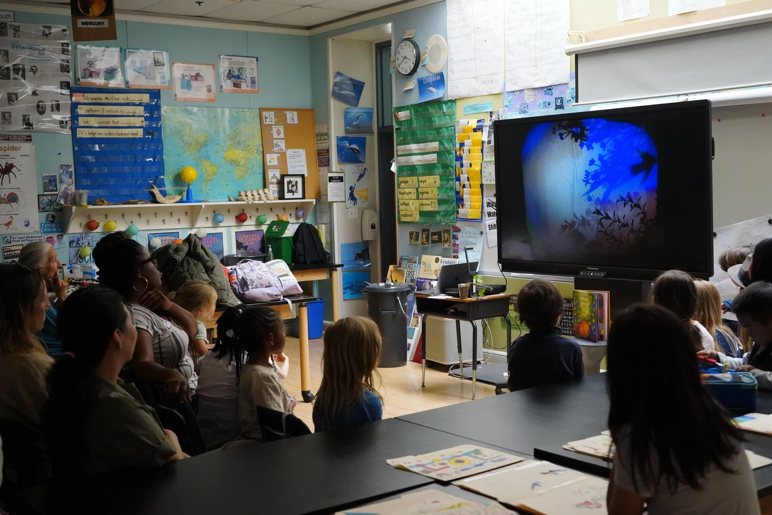 Classroom with children and adults watching a presentation on a large screen showing a forest scene with trees and birds.