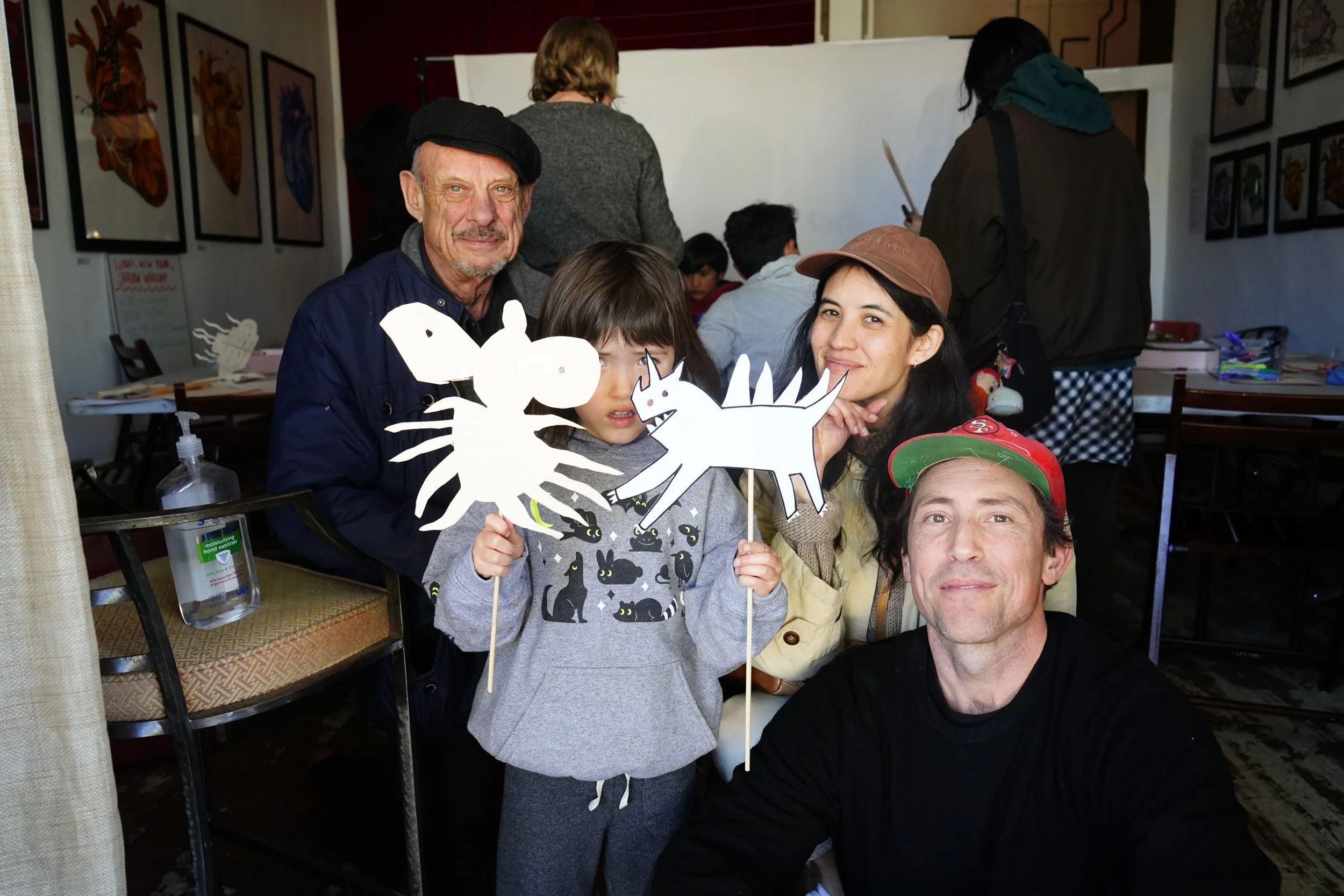 Group of five people, including a kid holding paper cutouts of animals, smiling in a decorated indoor space with artwork on the walls.