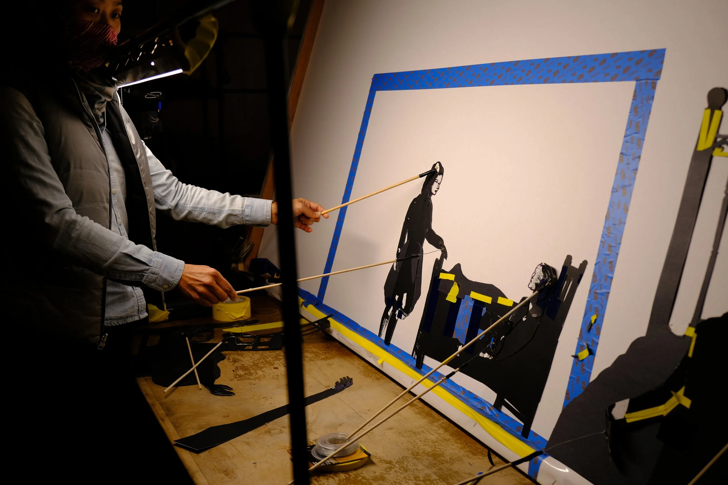 An artist creating a mixed media collage artwork on a large white canvas, using black paper cutouts of human figures and tape, with tools and supplies on a wooden table.