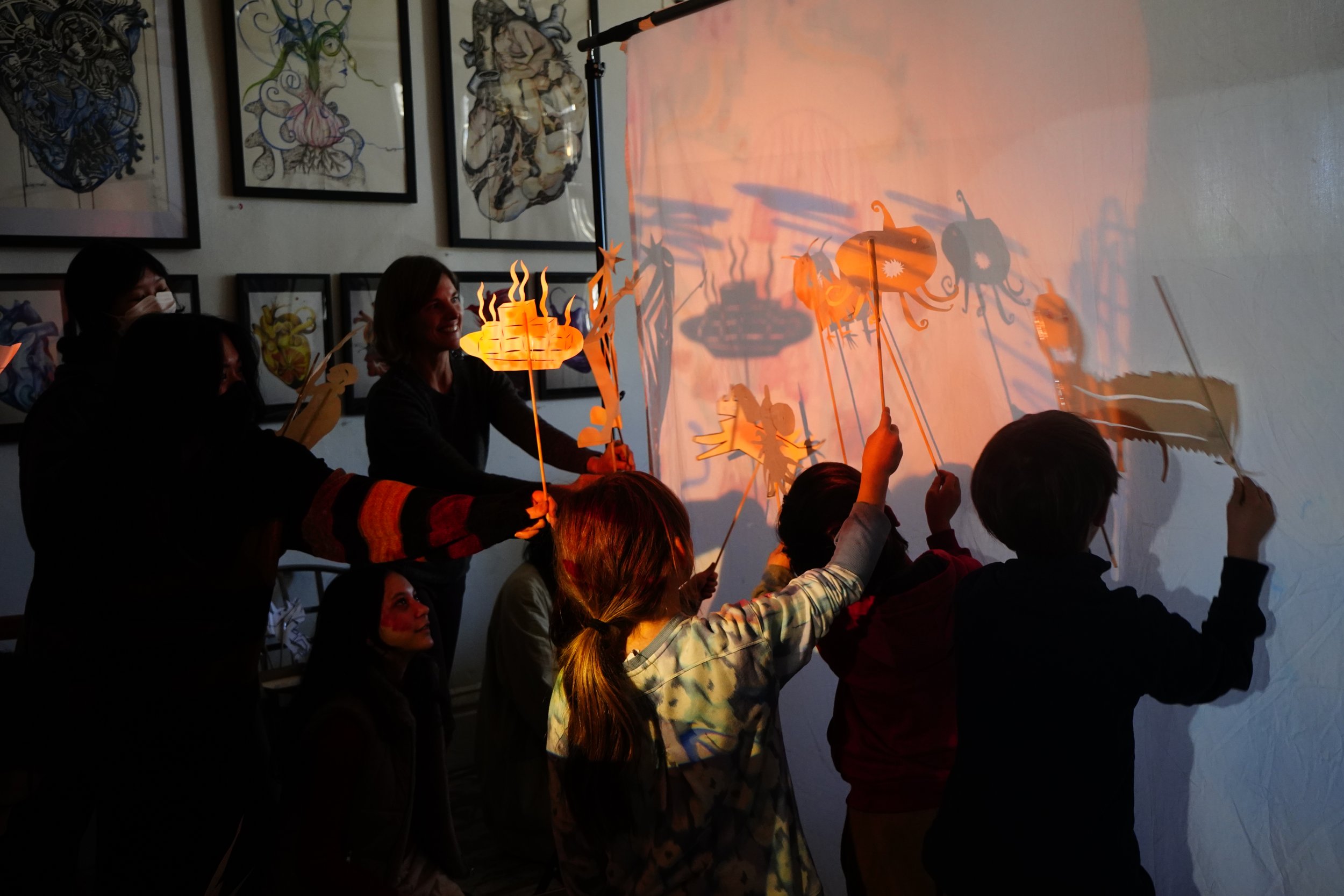 People participating in a shadow puppet workshop with lanterns, creating shadow puppets on a wall using sticks and lantern lights.
