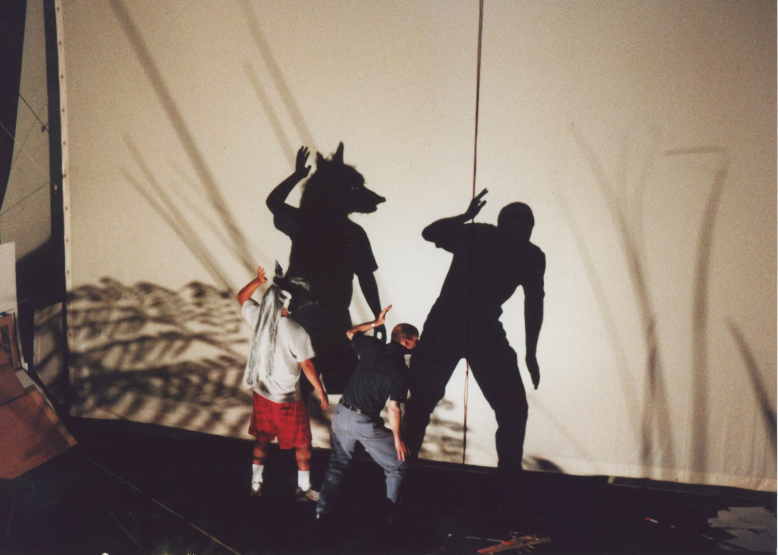Two people are standing in front of a white backdrop, creating shadows that resemble a woman with a wolf's head and a man with a raised arm. The shadows are projected onto the backdrop, creating an artistic shadow play.