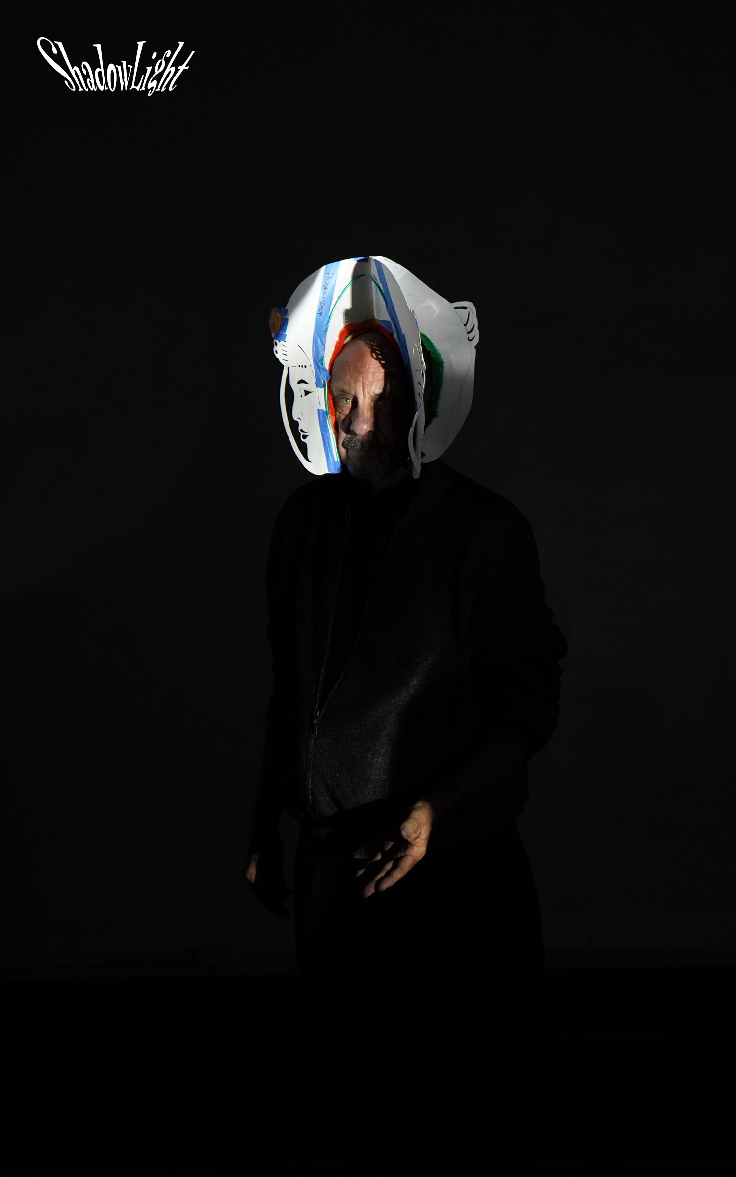 A person wearing a black jacket and gloves stands in a dark environment, with a shadowy face illuminated softly. They are wearing a large, artistic, and colorful headpiece resembling a paper sculpture or mask, with the word "ShadowLight" written in white cursive text in the top left corner.
