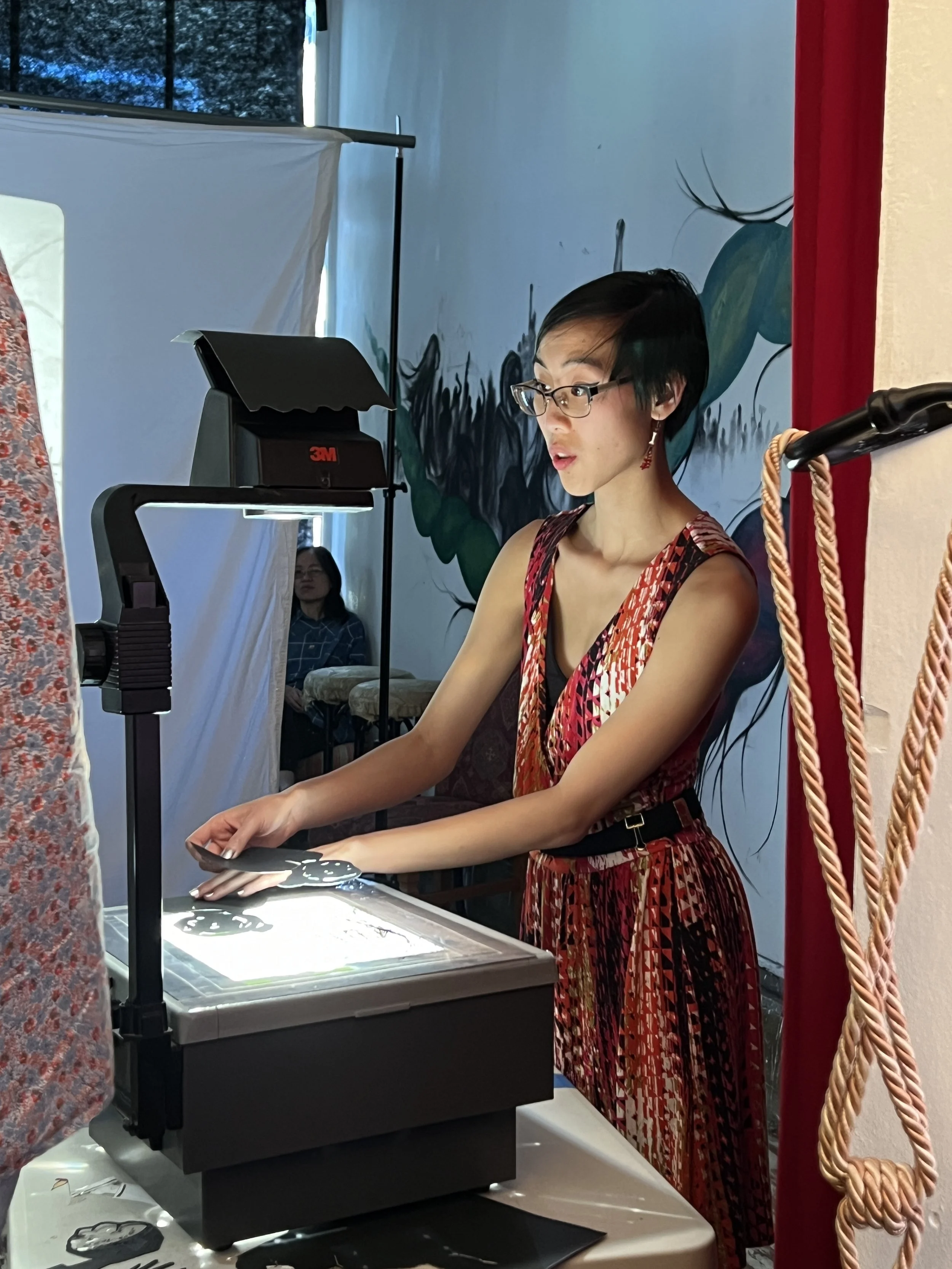 A woman with short black hair, glasses, and wearing a colorful, patterned dress operating a shadow puppet projection device. She stands next to a projection screen with a dark background, likely preparing or displaying a shadow puppet performance.