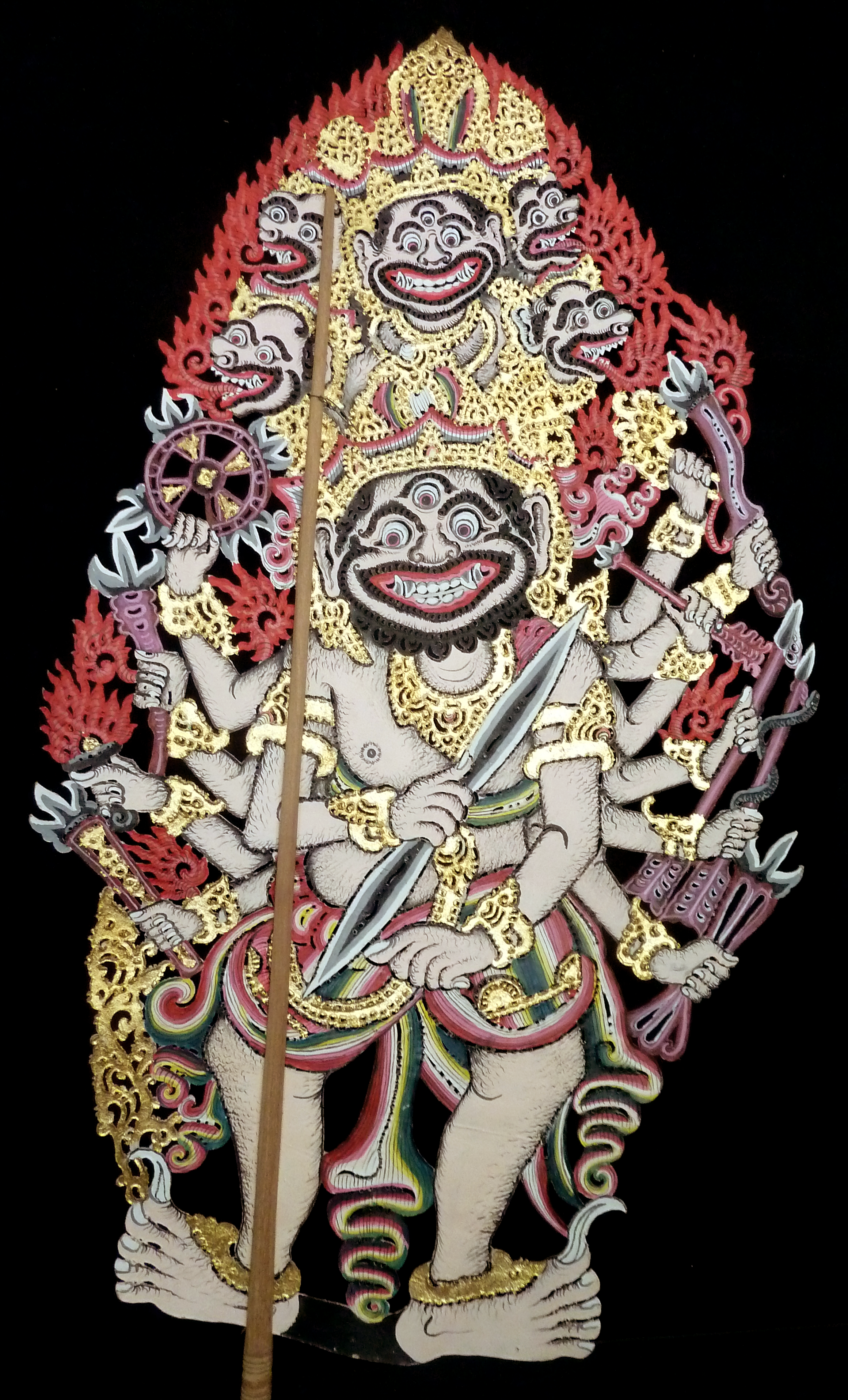 Colorful traditional Batak tor Tor puppet with multiple faces, adorned with gold details, holding weapons, against a black background.