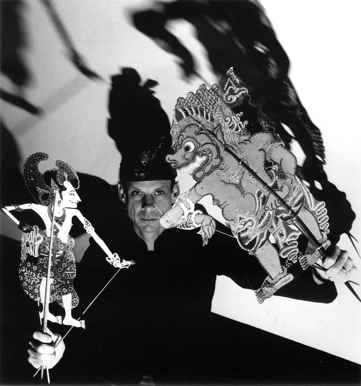 A person holding traditional puppets, including a figure wielding a spear and an armored beast, with shadows cast on the wall behind.