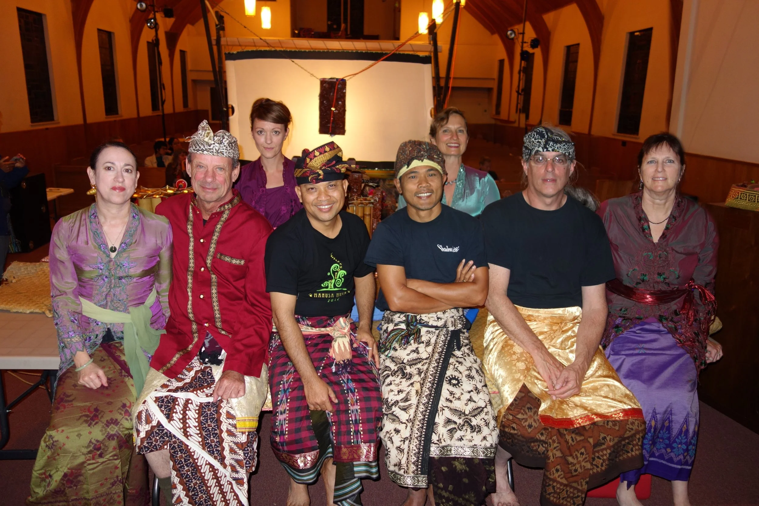 Group of nine people dressed in traditional Balinese attire, seated and standing in a hall, smiling at the camera.