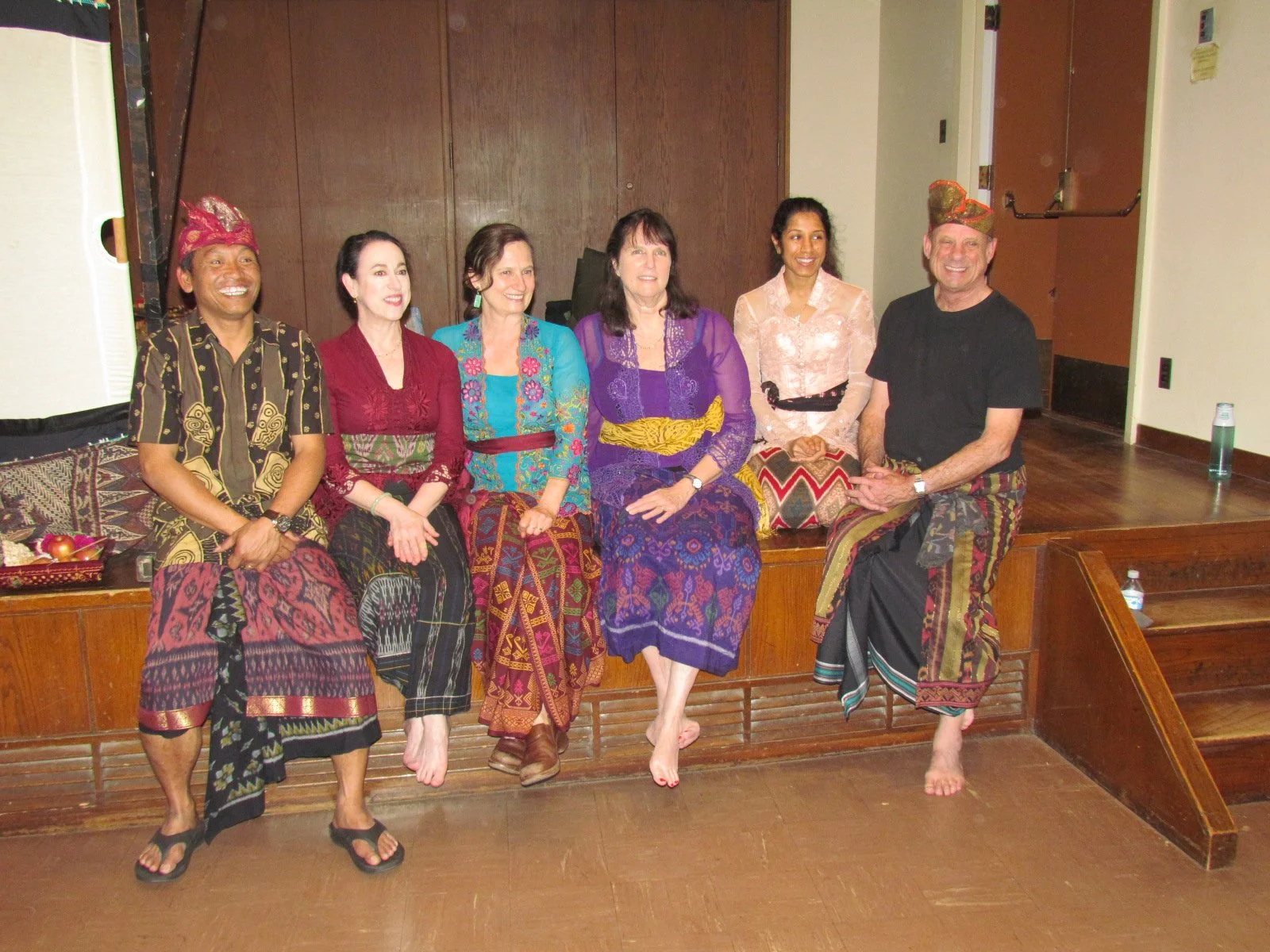 Group of six people sitting on a wooden bench, dressed in traditional Indonesian batik clothing, smiling for a photo in an indoor setting.