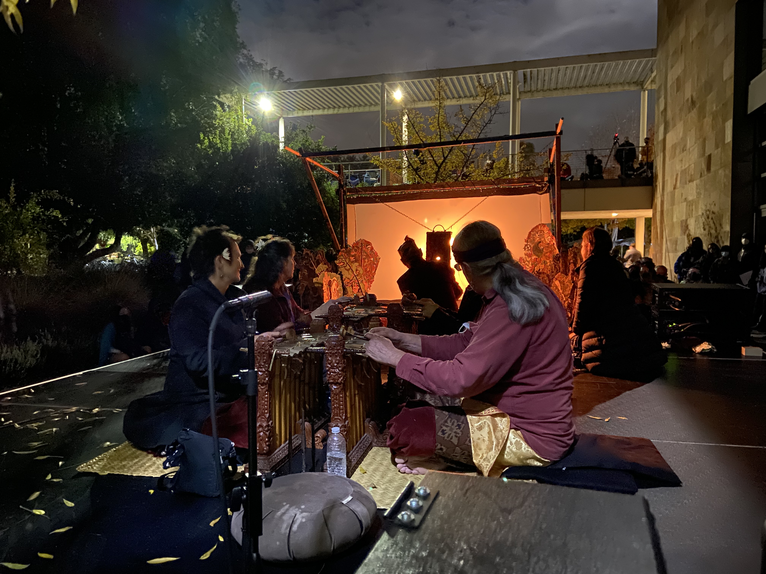 People sitting on the floor and preparing for a traditional shadow puppet performance at night, with a glowing orange screen and shadow puppets in the background.
