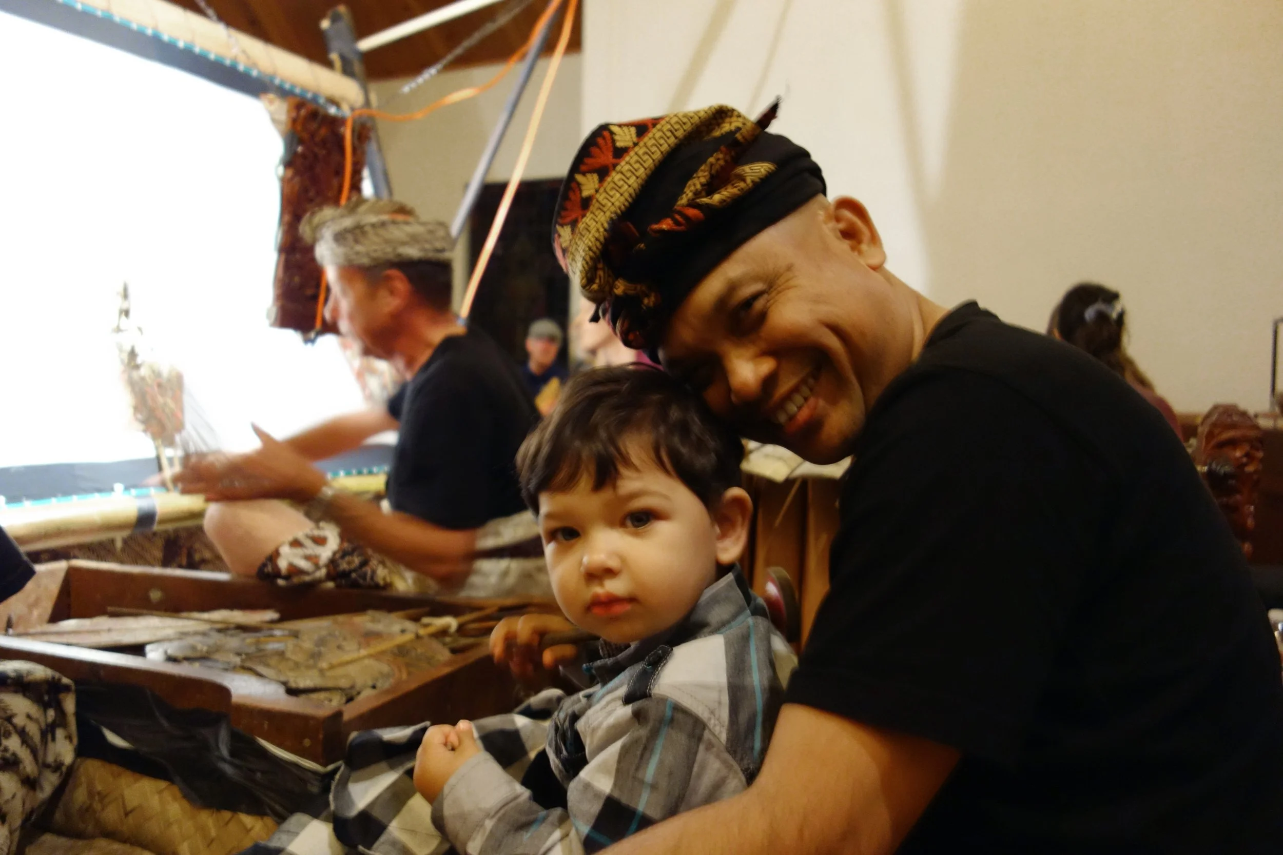 A man with a traditional patterned headwrap and a wide smile is holding a young boy with dark hair and a plaid shirt, both sitting at a table with craft supplies. In the background, a person is working on a craft project.