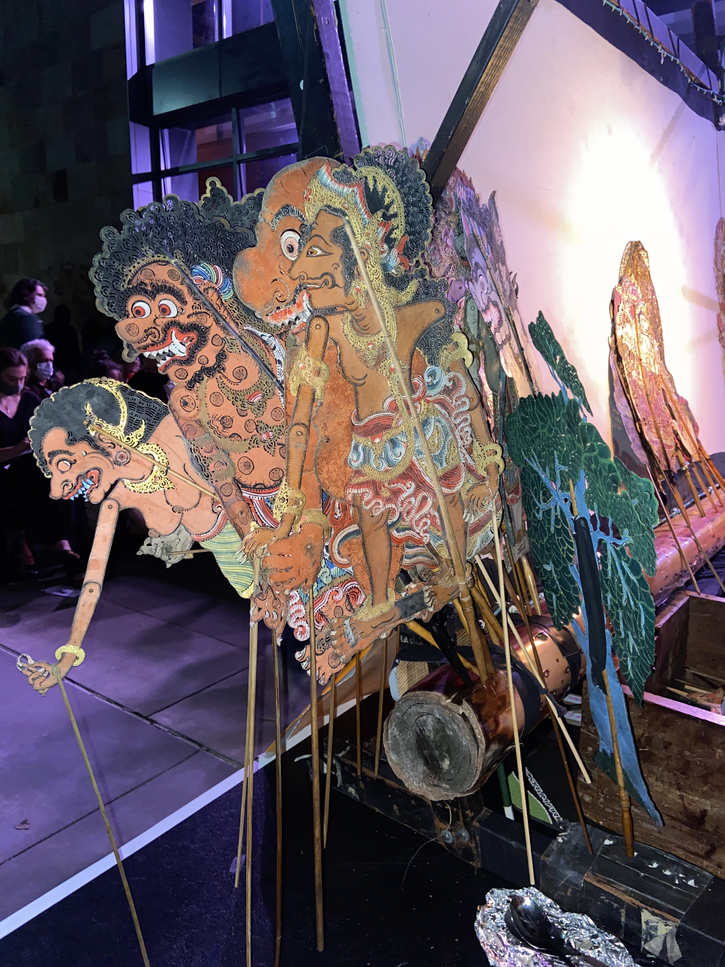 Colorful traditional Indonesian puppet puppets on display, depicting characters with exaggerated facial features, large eyes, and elaborate costumes, set against a background of mix of green, gold, and brown decorations.