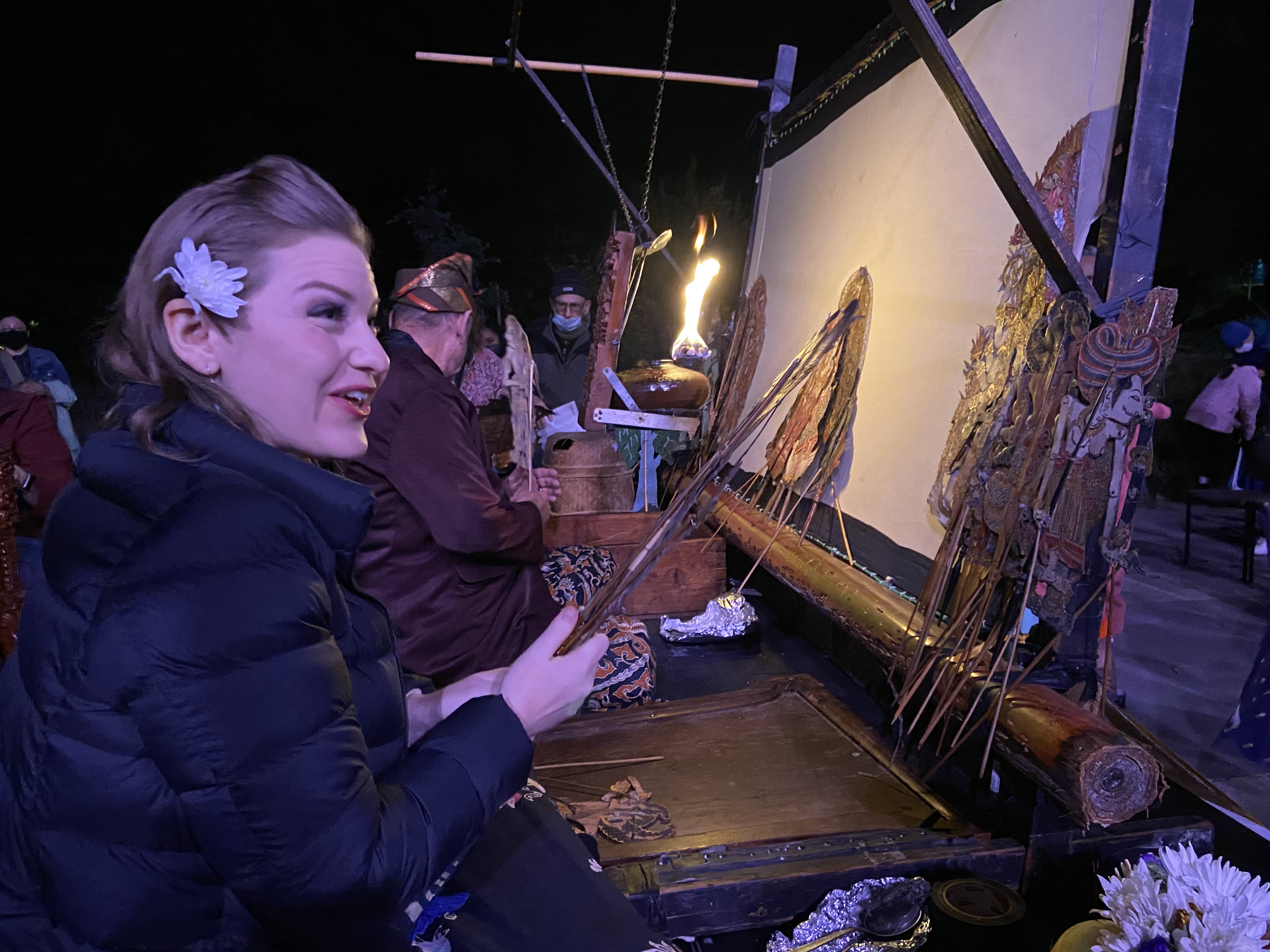 A woman with a white flower in her hair participating in a traditional puppetry performance, sitting beside an ornate wooden puppet stage at night.