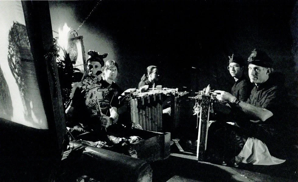 Group of performers, some wearing traditional Asian costumes, sitting at a low table with various objects in a dimly lit room, possibly preparing for a performance.