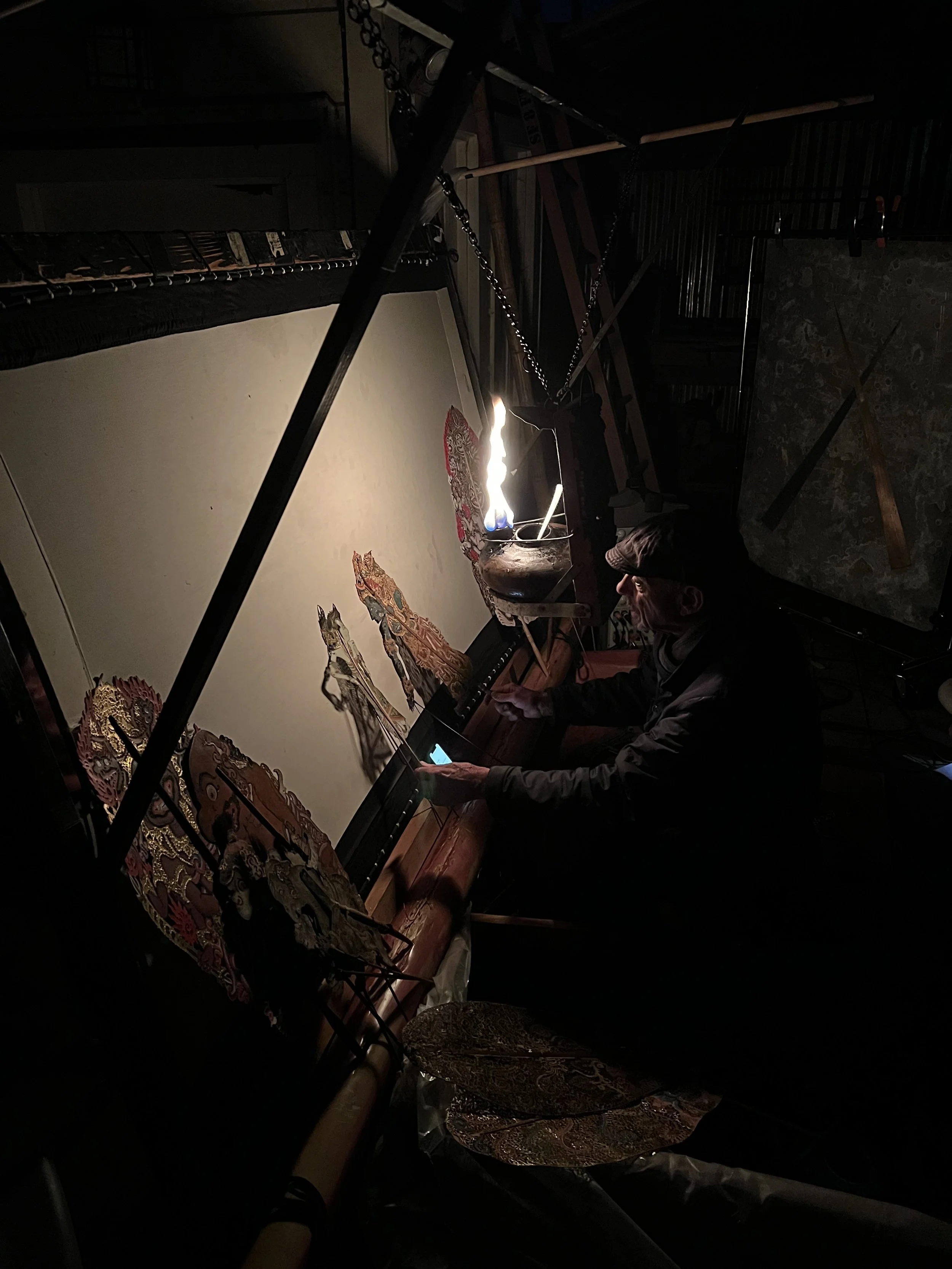A person working on a traditional shadow puppet show, manipulating puppets and using a fire-based light source behind a white screen.
