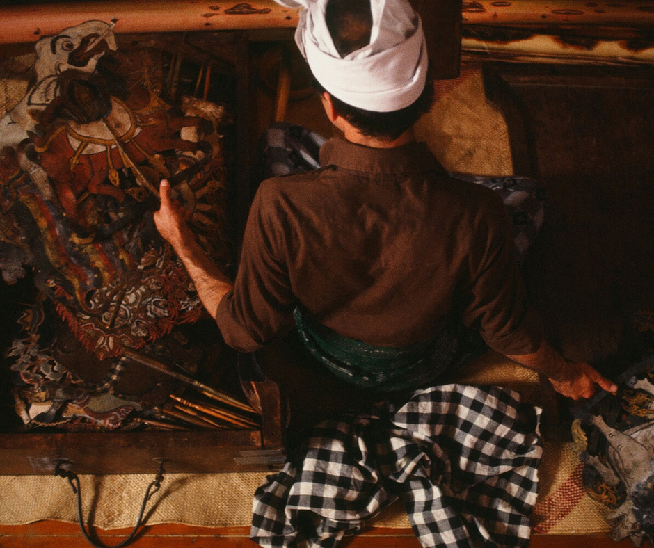 A person dressed as a chef sitting on a bench, viewed from above, with traditional textiles and art around, including a tiger painting.