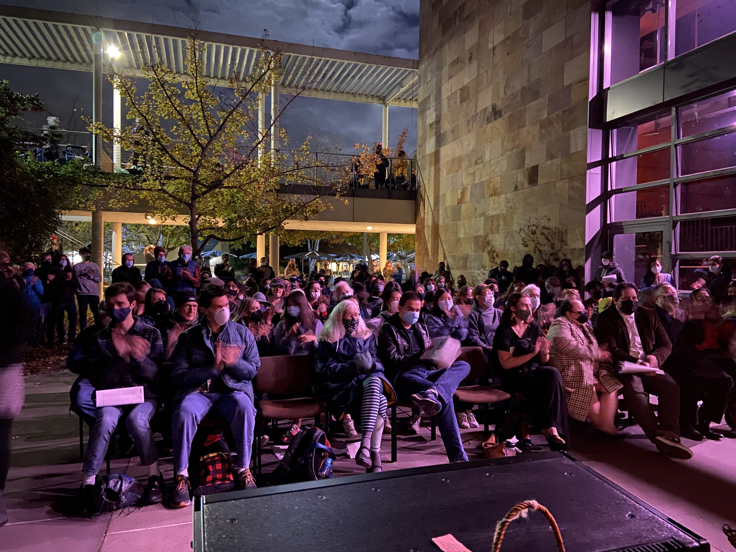 Audience wearing masks attending an outdoor event at night, seated and standing in front of a modern building with illuminated purple and pink lighting.