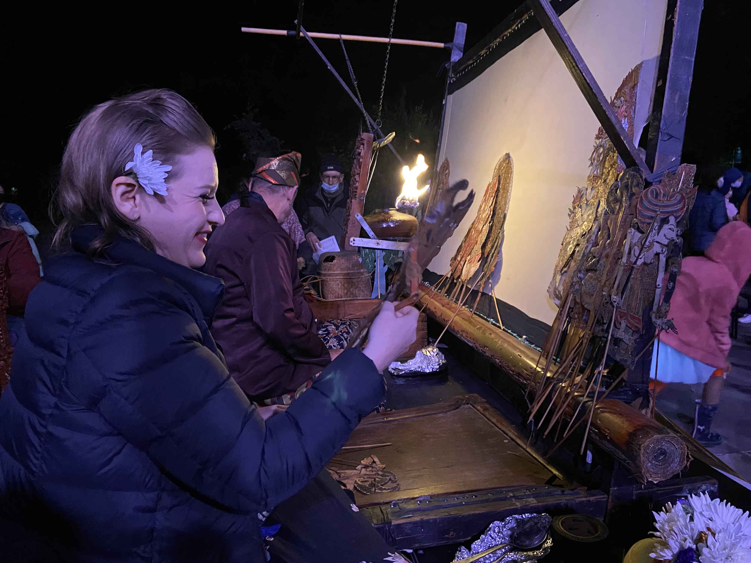 A woman with a white flower in her hair is smiling while participating in a traditional shadow puppet performance at night. She is holding a puppet and sitting at a table with various puppets and a light, with multiple people observing in the backgro