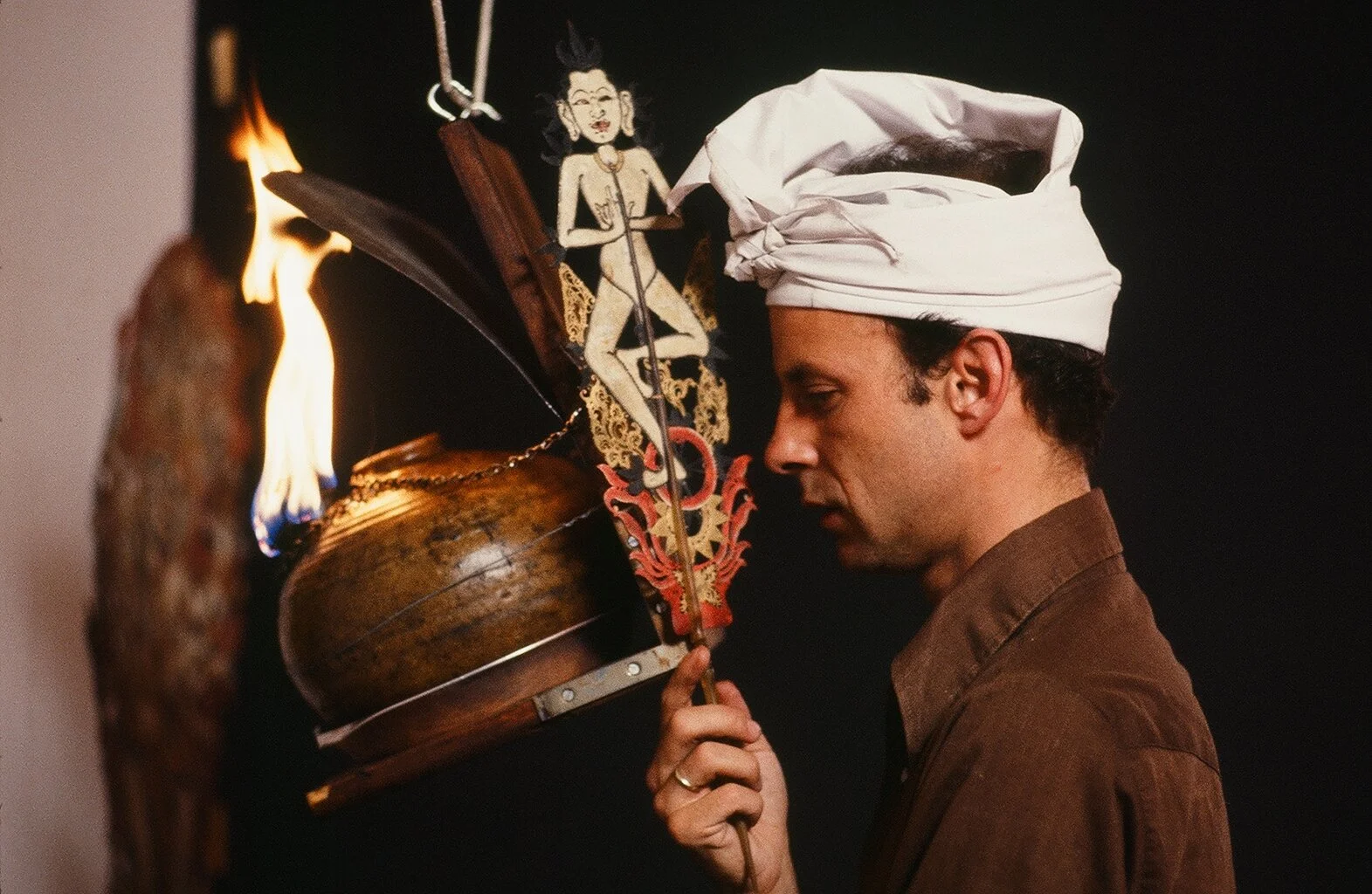 A man wearing a white chef hat, holding a torch with a burning flame, is facing a display featuring a puppet of a woman with black hair and a yellow dress, along with other decorative items.