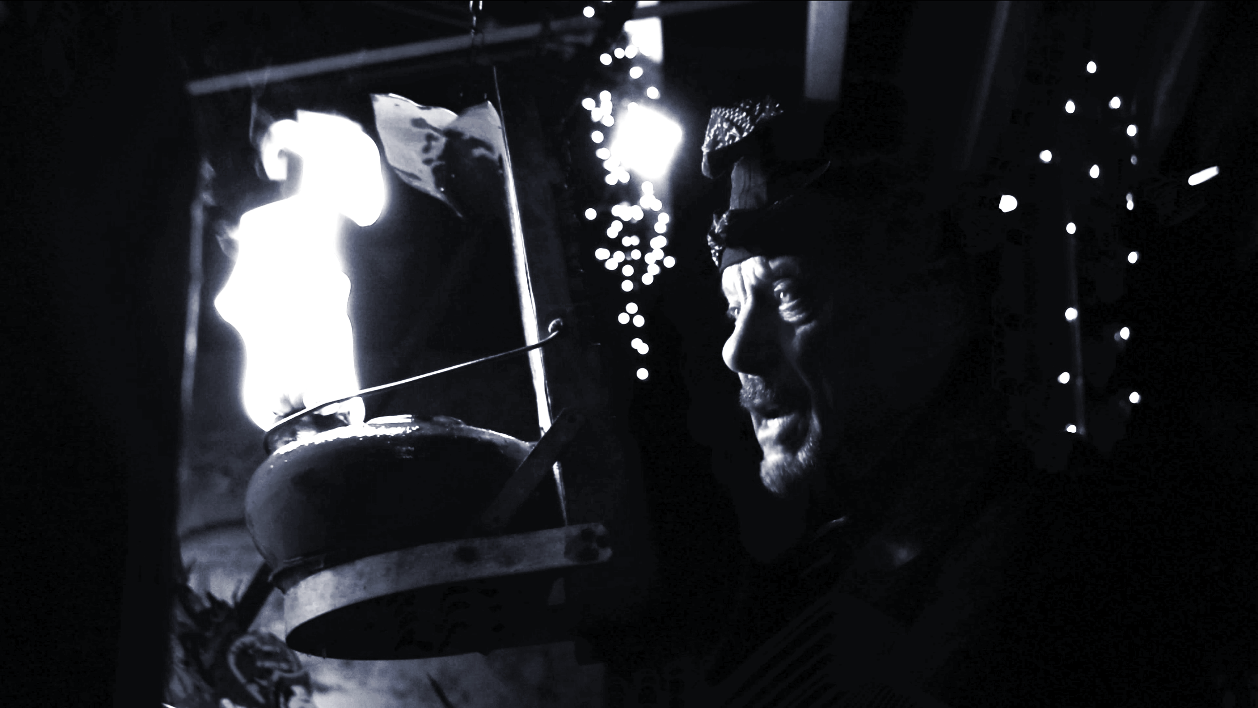 A person wearing a bandana is lighting a traditional oil lamp with a flame, surrounded by hanging string lights in a dark setting.