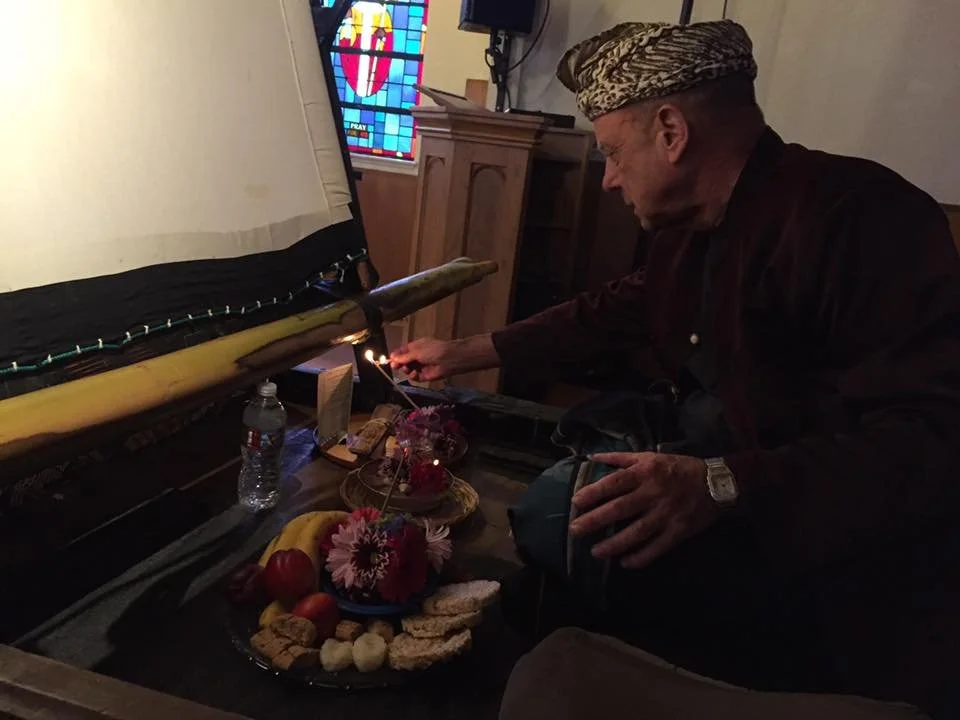 An elderly man with a headscarf lighting a candle near a large prayer scroll on a table with fruit, flowers, and snacks inside a church.