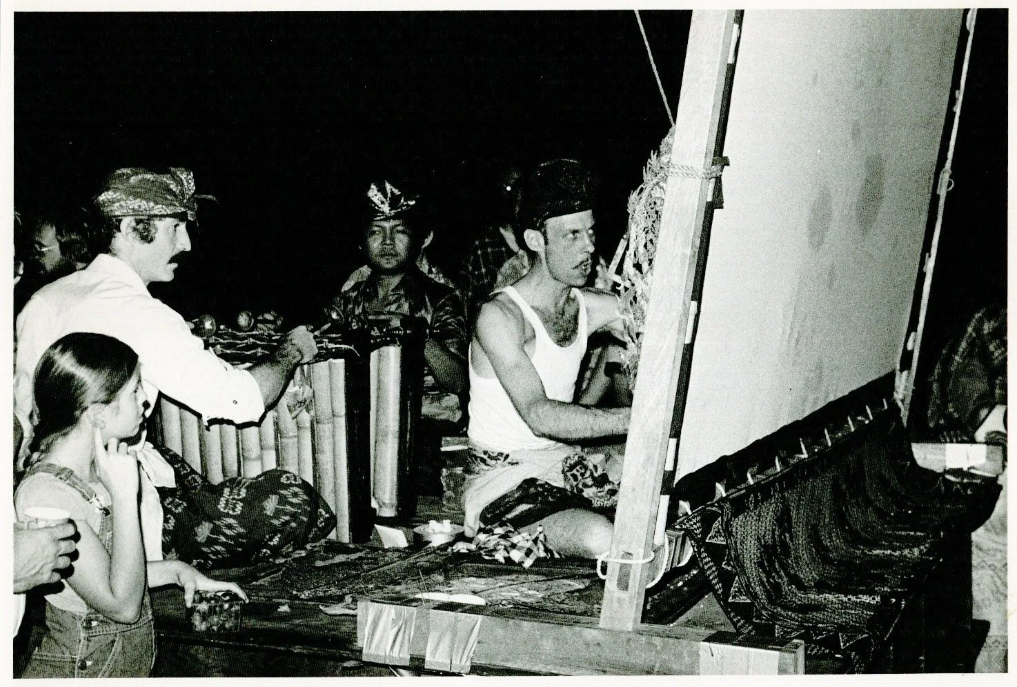 A group of men and a girl gathered around an artist painting on a canvas, with some wearing traditional Indonesian clothing and headgear, at night.