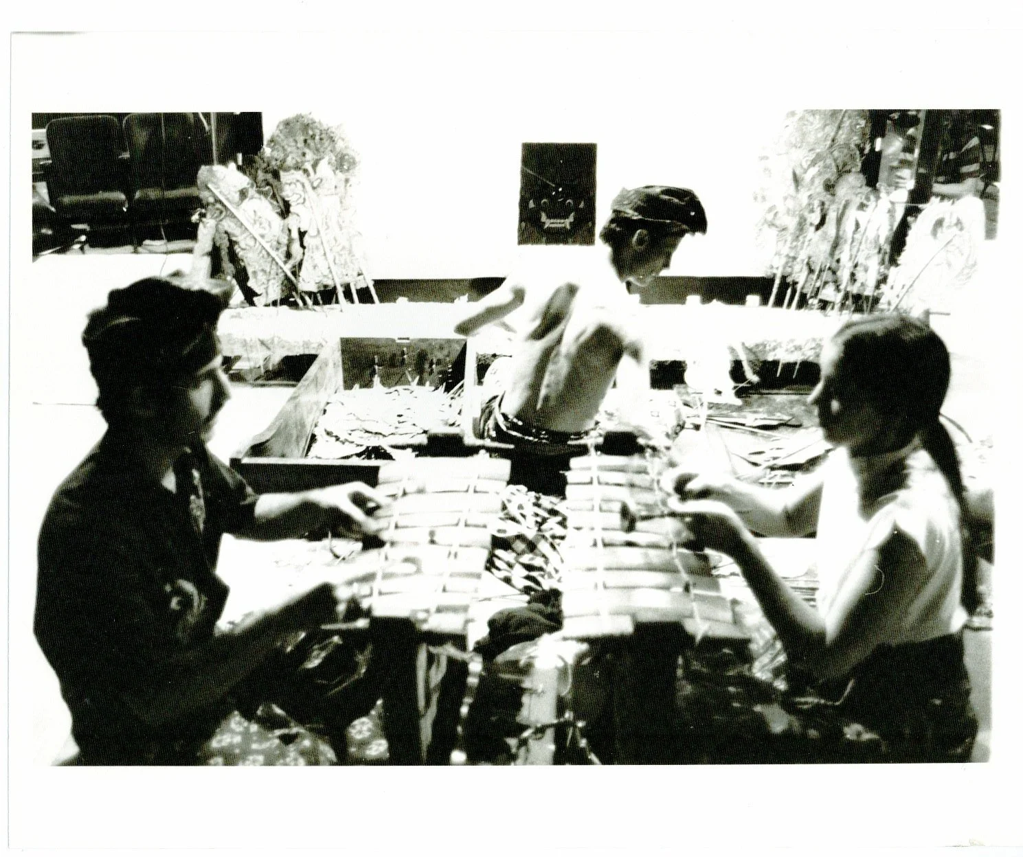 Three people sitting and one man standing in a room with artworks on the wall, possibly creating or examining art pieces.