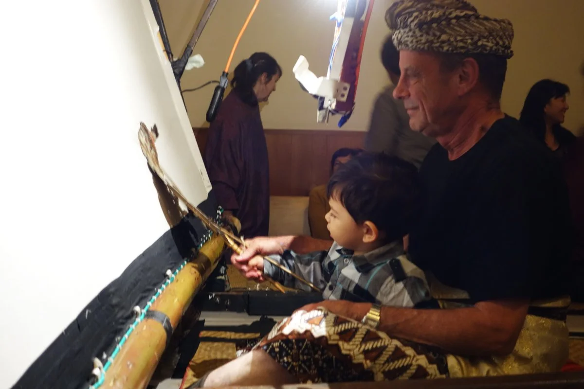 An elderly man wearing a traditional patterned headwrap and a black shirt helps a young boy, possibly his grandson, paint a large traditional drum together. They appear to be on a stage or in a performance space, with others watching and smiling in t