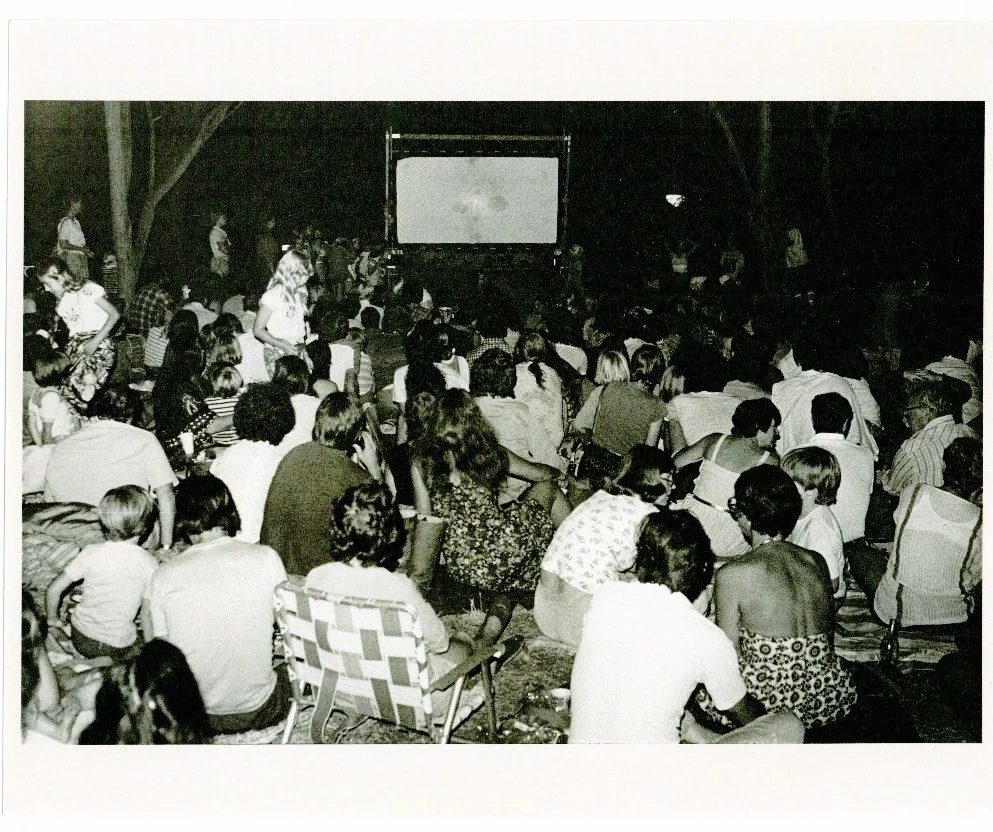 A large group of people seated outdoors at night, watching a movie on a big screen.