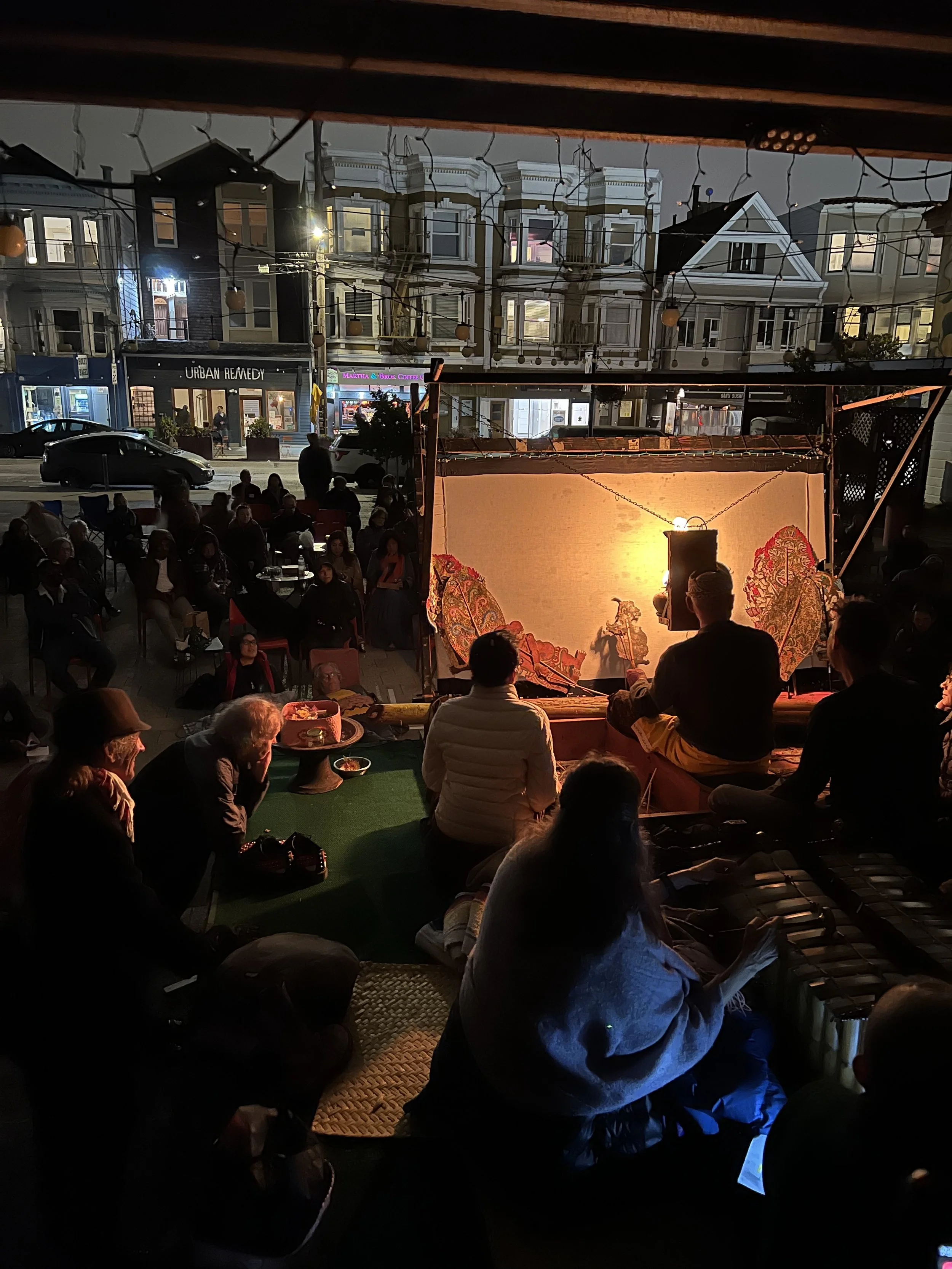 An outdoor puppet show at night with an audience seated on the ground and in chairs, illuminated by warm lighting with houses and shops across the street in the background.