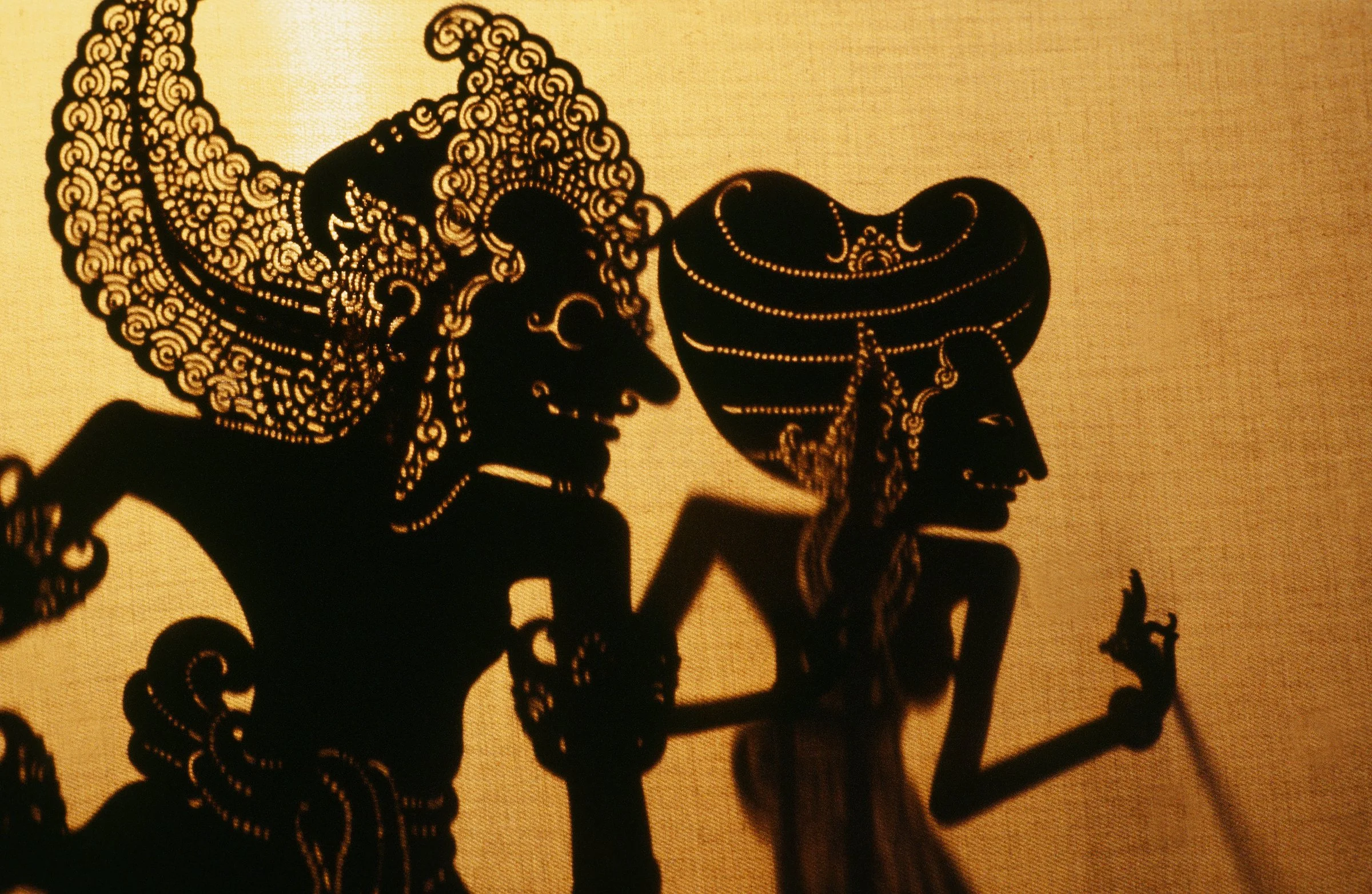 Silhouette of two traditional Indian shadow puppets with intricate details, depicting a man and a woman in traditional attire, against a warm light background.