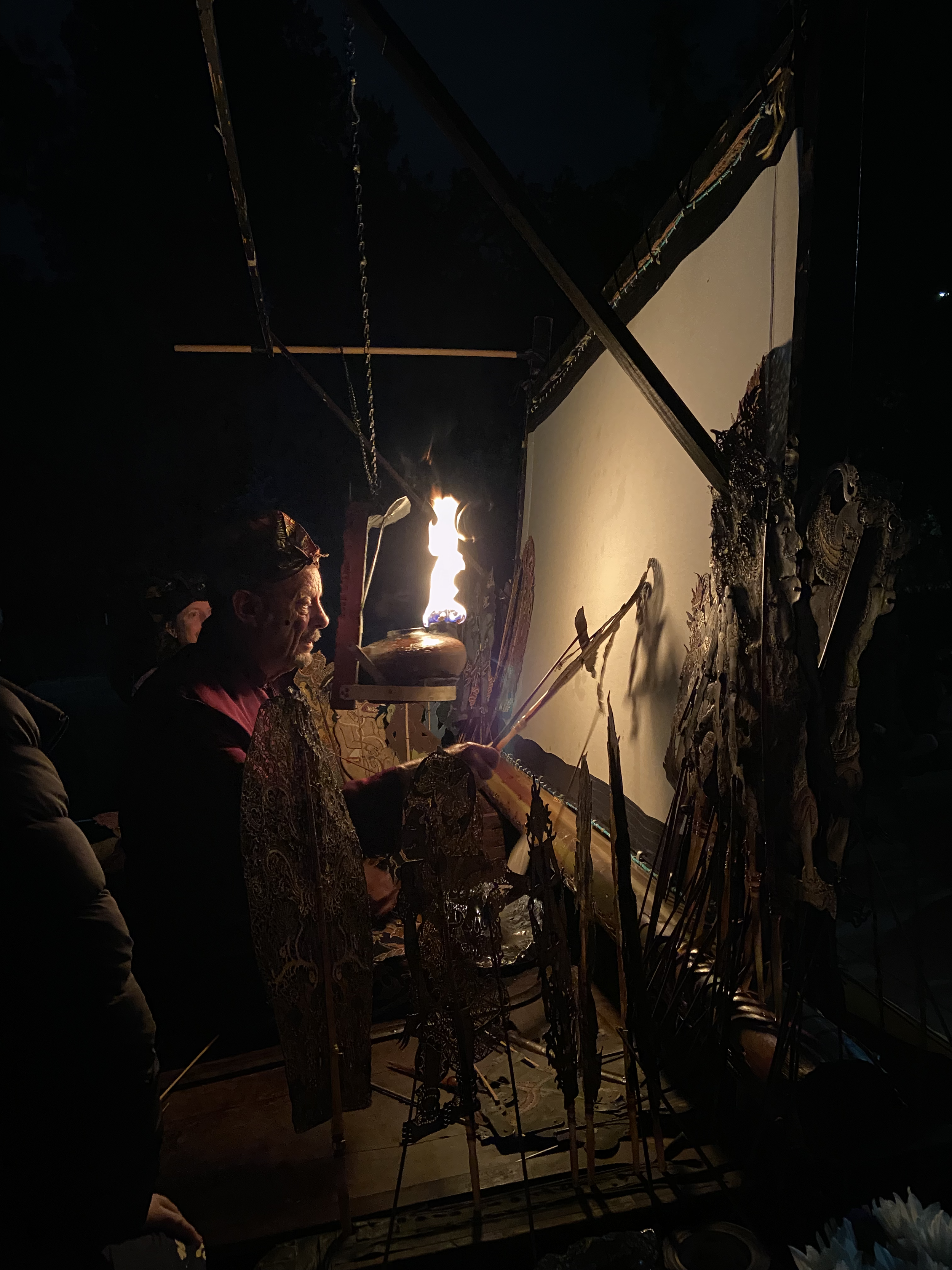 A person performing a traditional fire ritual outdoors at night, with various ritual objects and a large blank screen or canvas behind them.