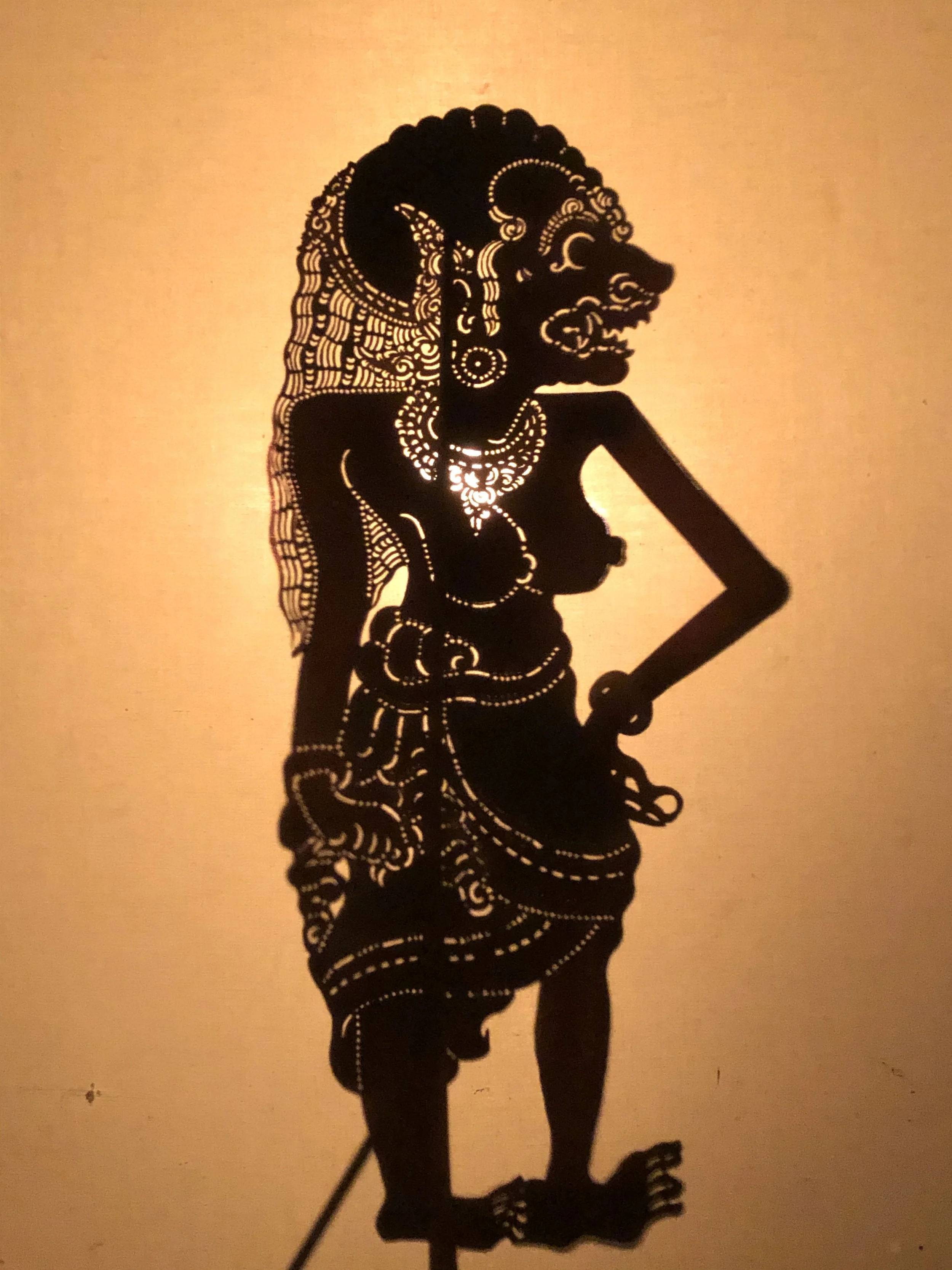 Silhouette of a mythological figure with elaborate jewelry and adornments, standing with one hand on its hip, against a warm light background.